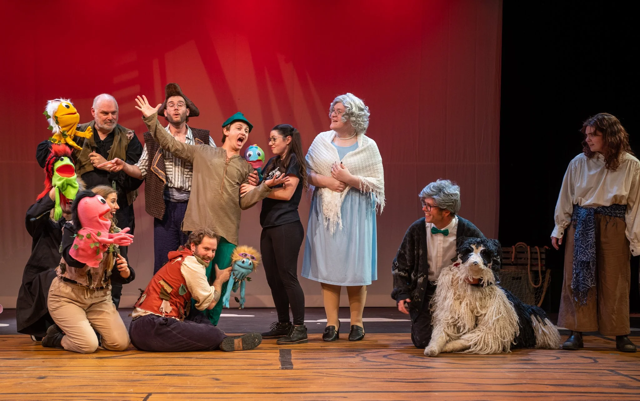 Group of performers on stage dressed as characters and puppets, with some in costumes and others in casual clothing. A large puppet is being held by a performer, and a large dog puppet is sitting in front. The scene appears to be from a theatrical pr