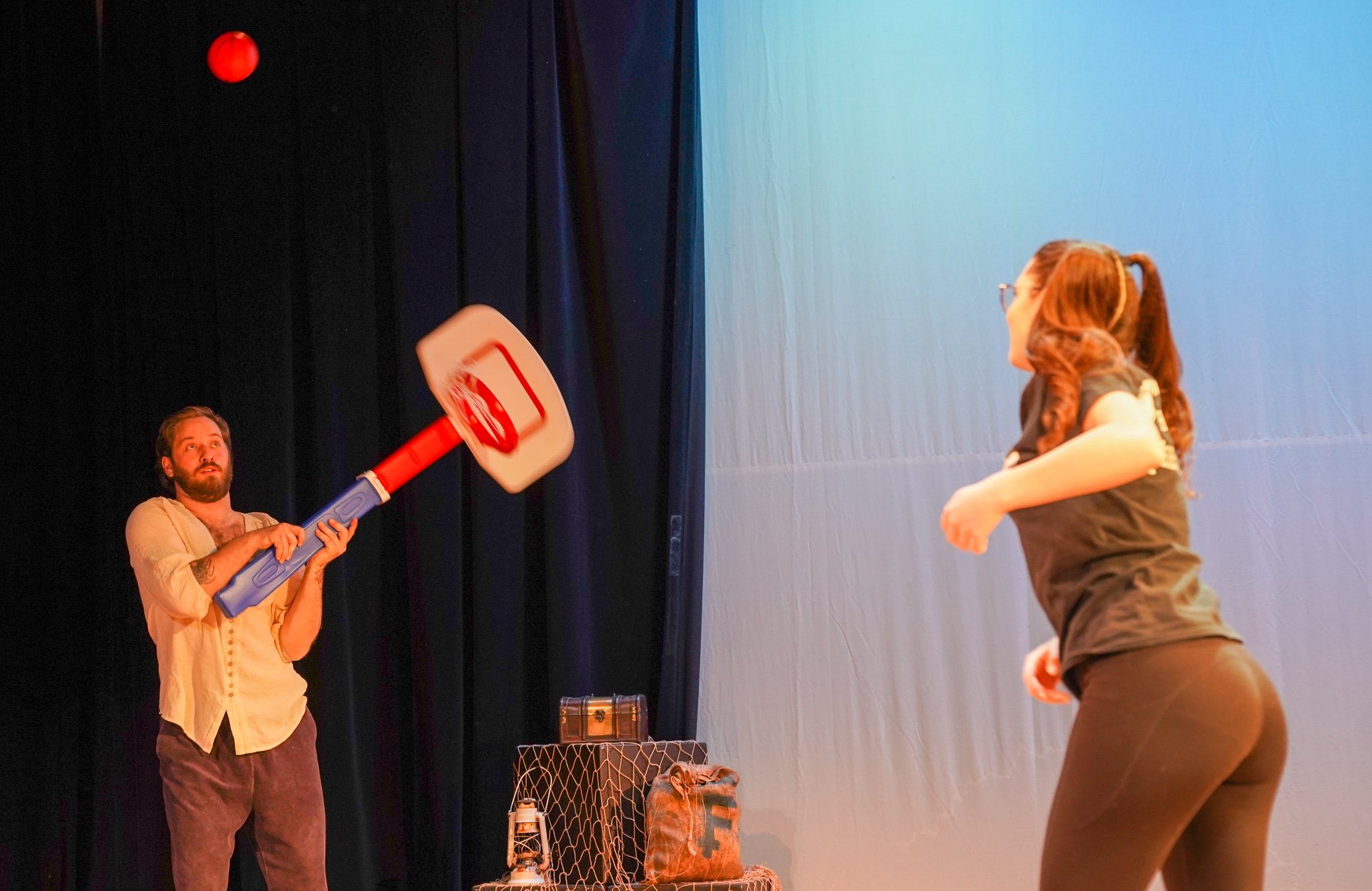 A man with a beard is holding a toy basketball hoop with a ball about to be shot into the hoop. A woman with glasses and long hair is standing in front facing the man, with her arm slightly raised. The scene appears to be on a stage or in a room with