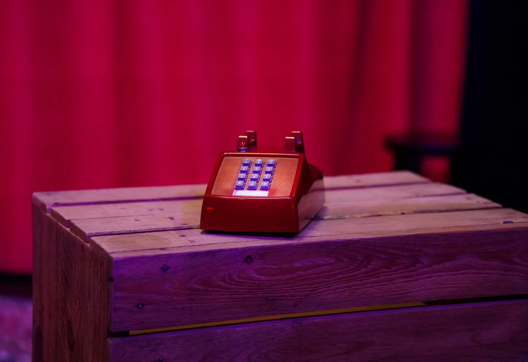 A vintage red rotary telephone placed on a wooden table with red curtains in the background.