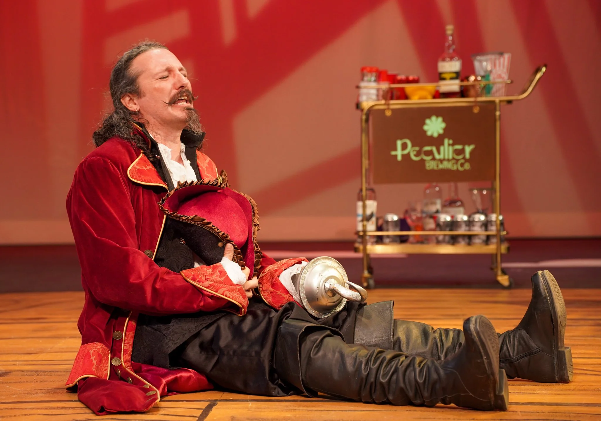 A man dressed as a clown laughing while sitting on a wooden stage, with a cart of snacks and a sign that reads "Peculiar Beverages Co." in the background.