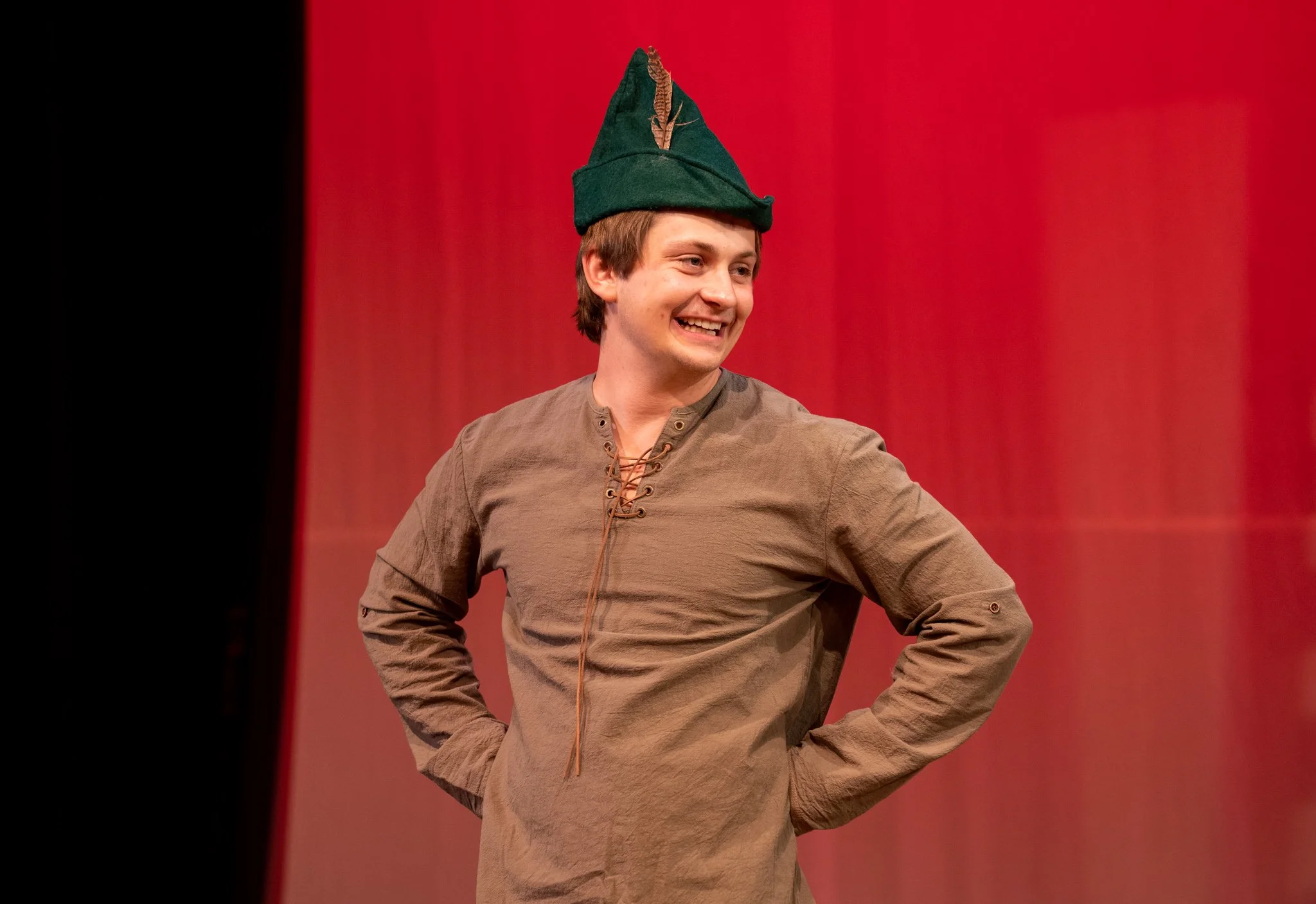 A young man wearing a green hat with a brown feather, smiling, standing in front of a red background.