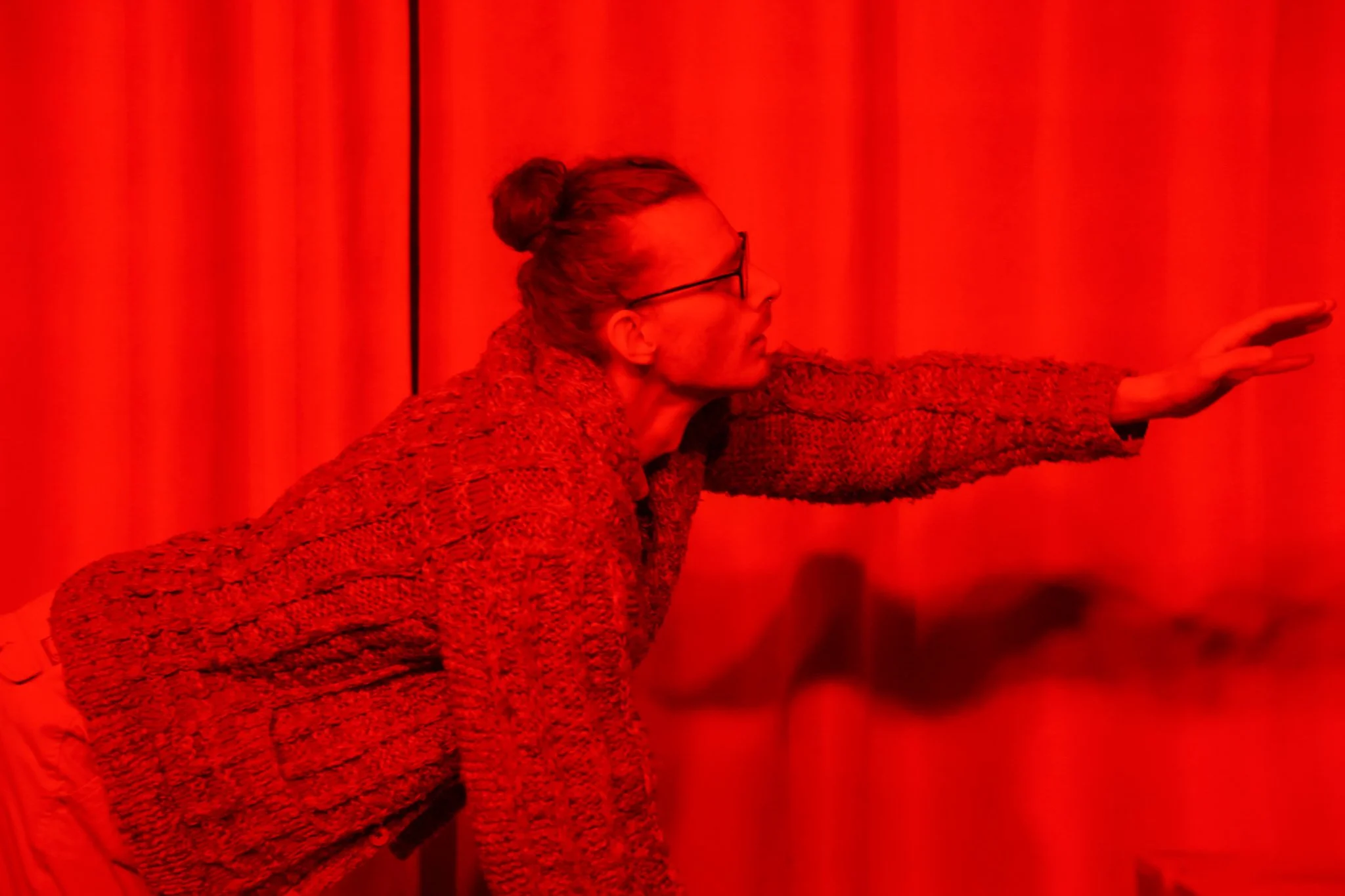 A person wearing glasses and a patterned sweater extending their arm forward, set against a red background with curtains.
