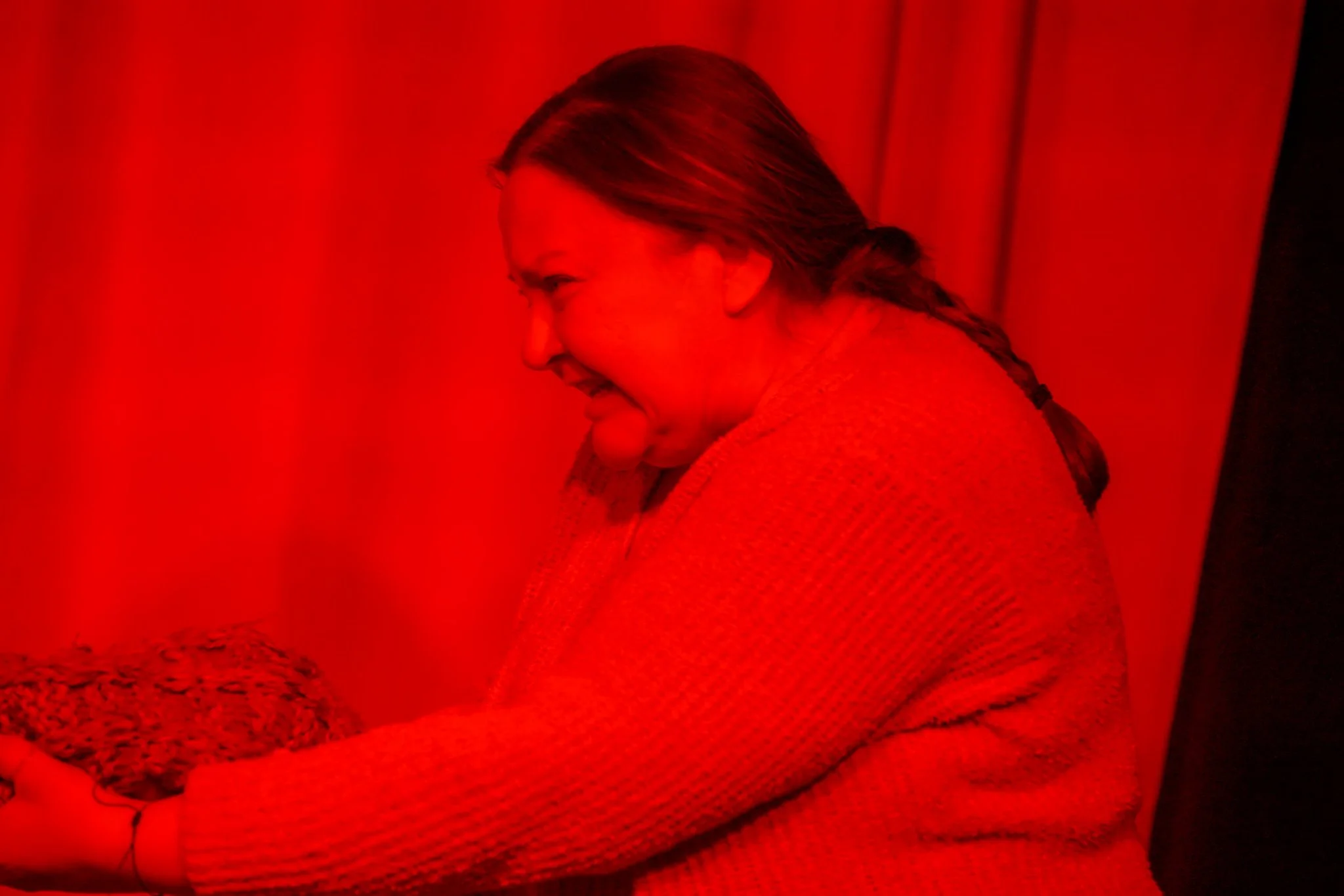 A person with long dark hair in a braid, leaning towards a wall, illuminated by a red light, wearing a sweater, with a part of their face smiling.