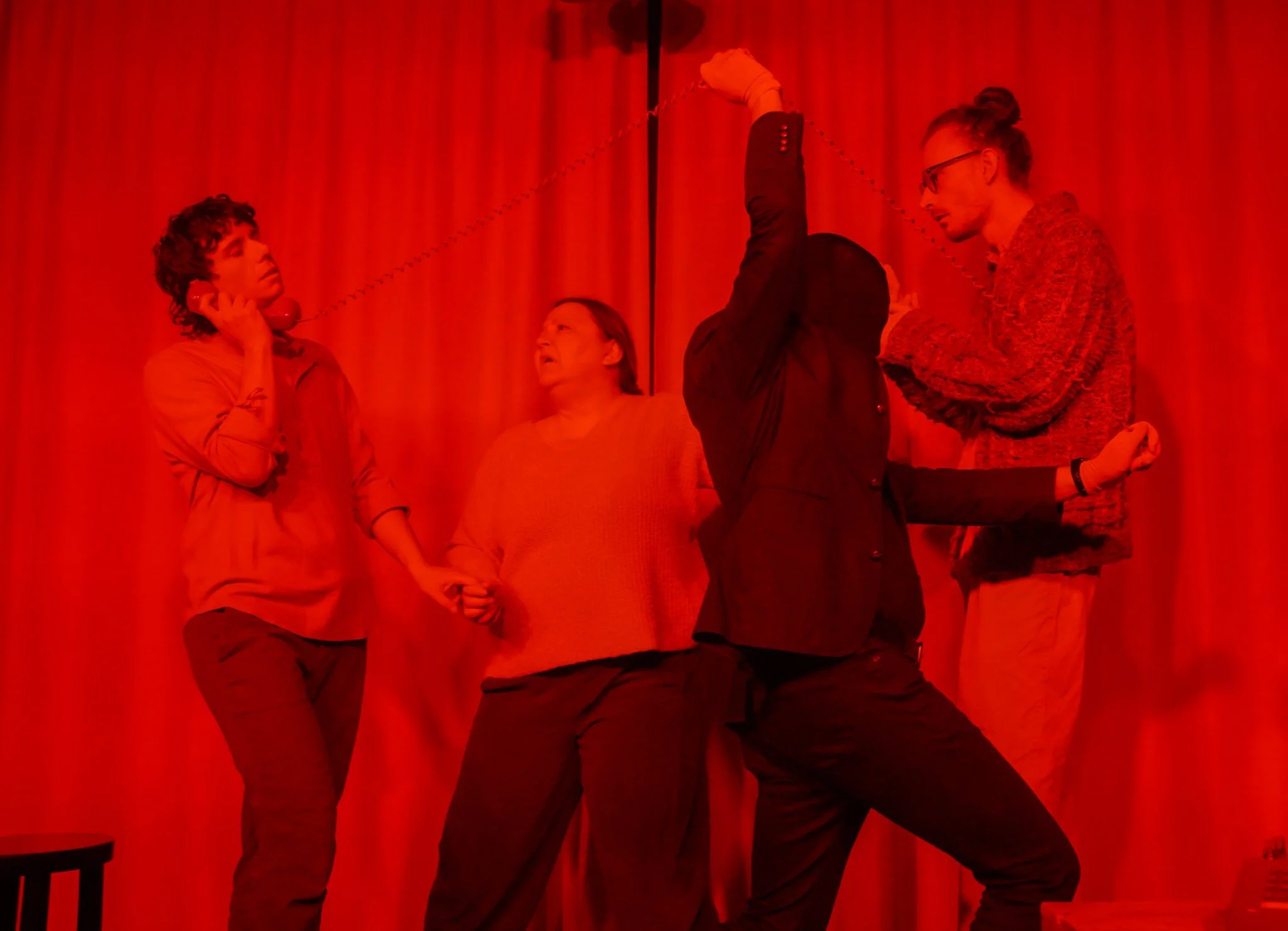 Four people are posing against a red curtain background. One person is holding a vintage red telephone receiver to their ear, while another person is raising their arm with a chain attached to a man holding the chain. The group appears to be engaging
