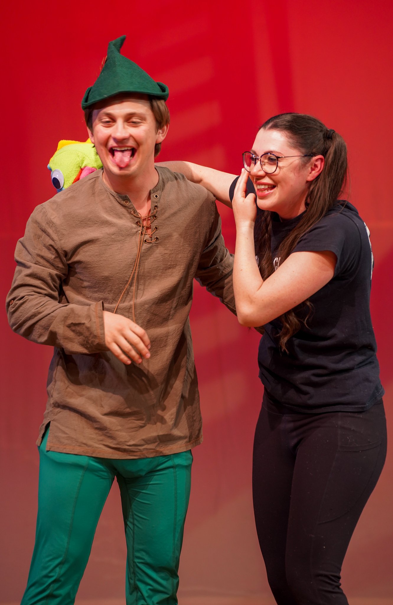 Two friends laughing and smiling, one wearing a green elf hat with a feather and a plush toy on his shoulder, the other wearing glasses and a black T-shirt, set against a red background.