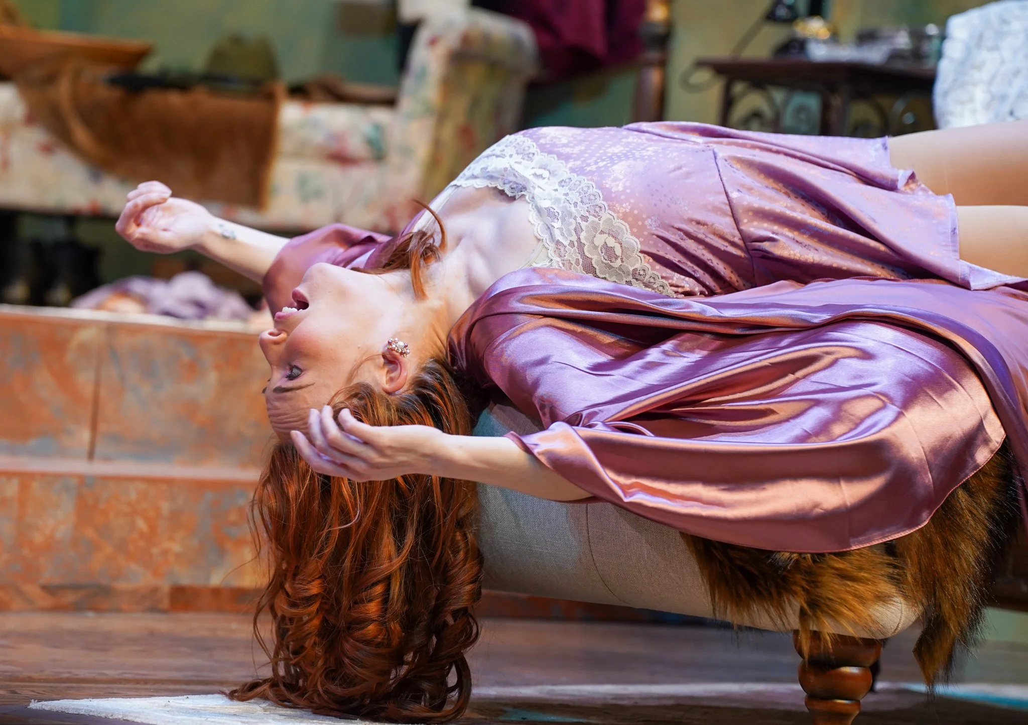 A woman with red hair is lying on the floor with her head hanging upside down over a chair. She appears to be laughing or smiling and is holding her hair with one hand. She is wearing a shiny pinkish-purple dress with lace details.