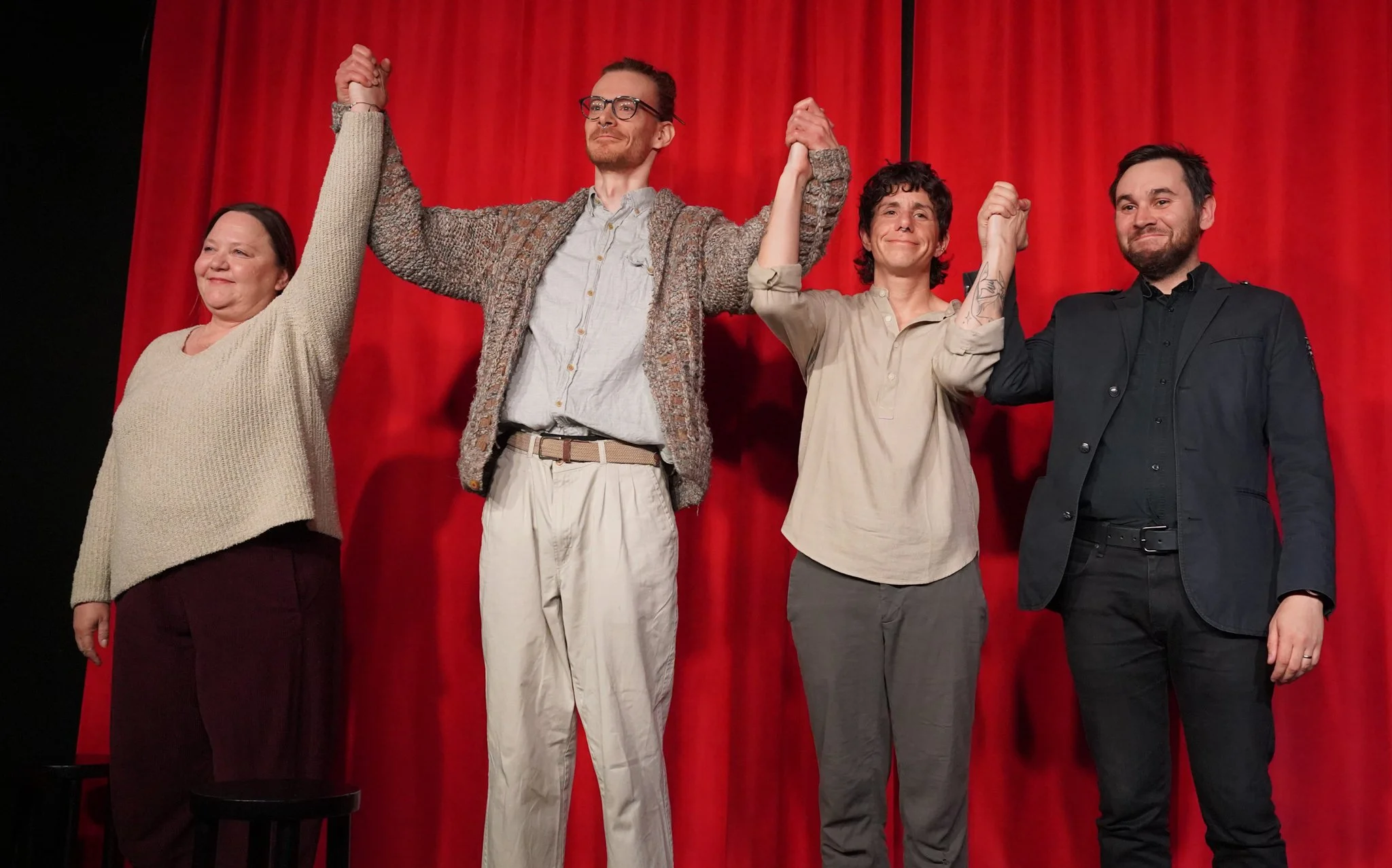 Four people standing in front of a red curtain, holding hands up in the air, smiling, and celebrating.