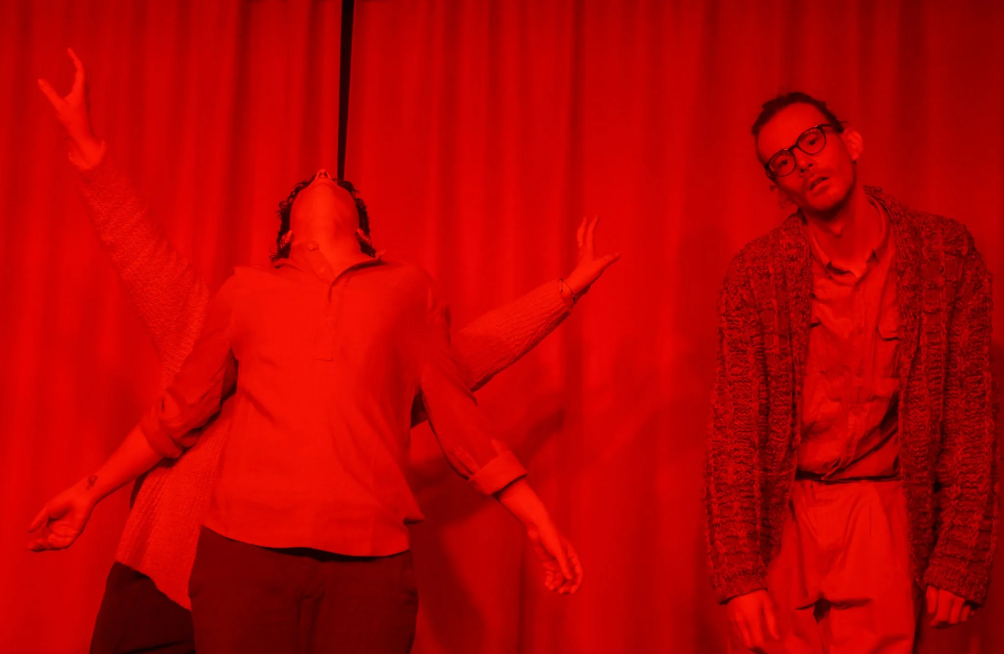 Two people stand against a red curtain background. One person is wearing a patterned jacket and glasses, while the other is wearing a jacket and is stretching outward with one arm. The scene has a red lighting or filter.