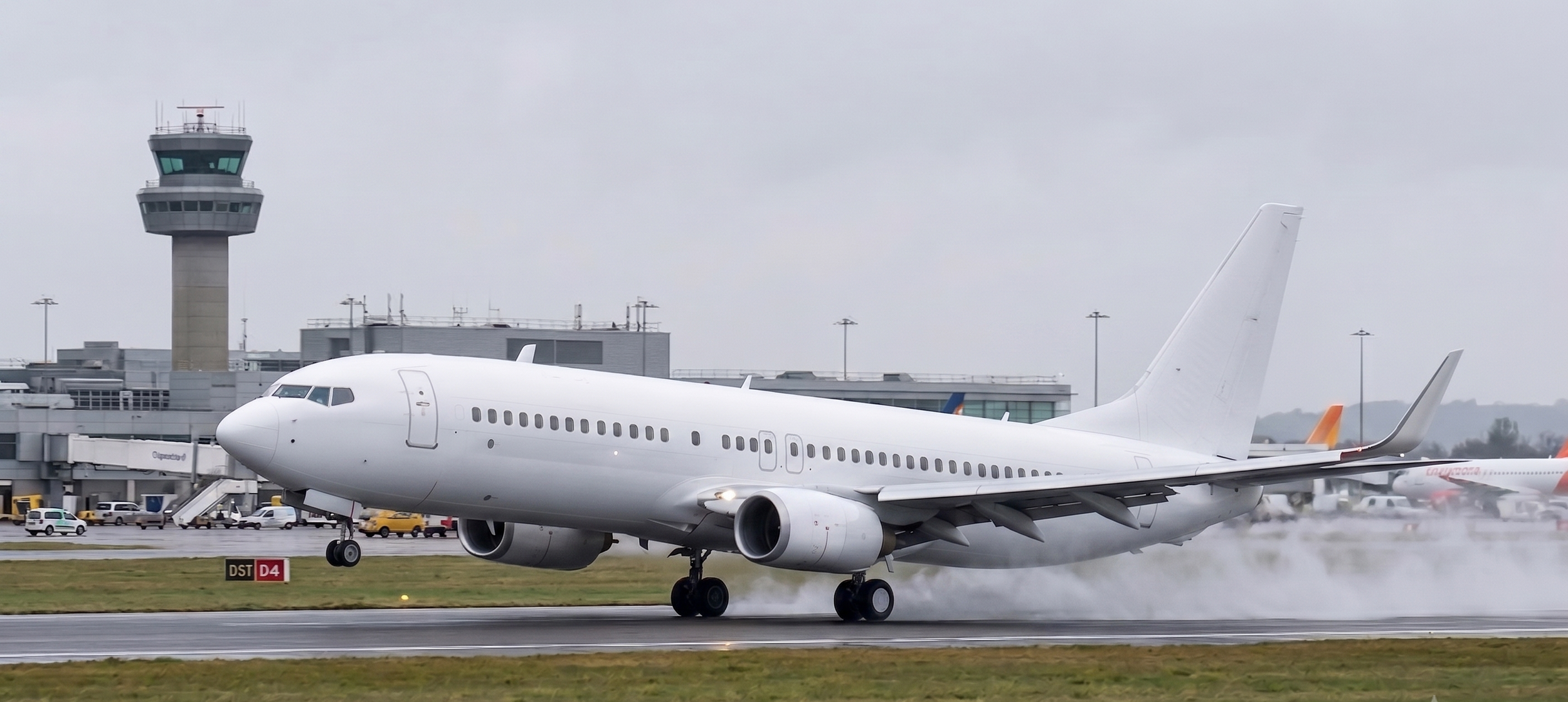 A white airplane landing on a wet runway at an airport, with an airport control tower and terminal building in the background.