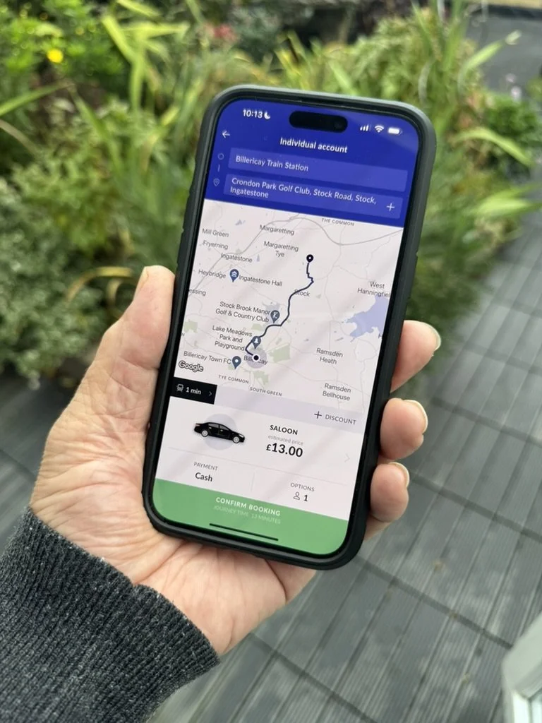 A person holding a smartphone with a ride-sharing app open, showing a route map from Billericay Train Station to Crondon Park Golf Club, with a black car icon, fare of £13, payment method as cash, and a confirmation button at the bottom.