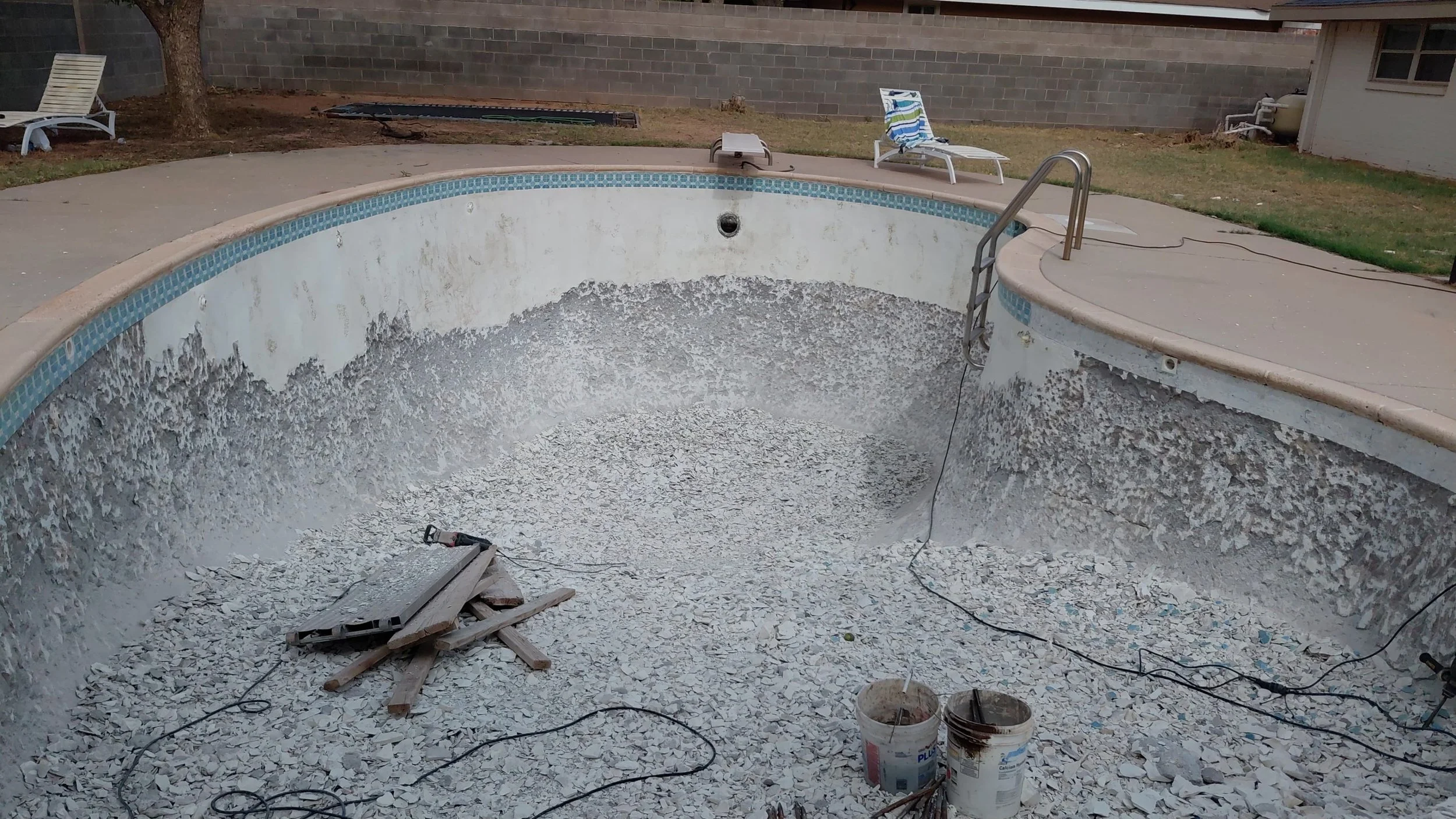 Empty swimming pool undergoing renovation, with debris and construction tools inside and around it, including buckets, wood planks, and electric cords; backyard with lawn, lounge chairs, and a brick wall in the background.