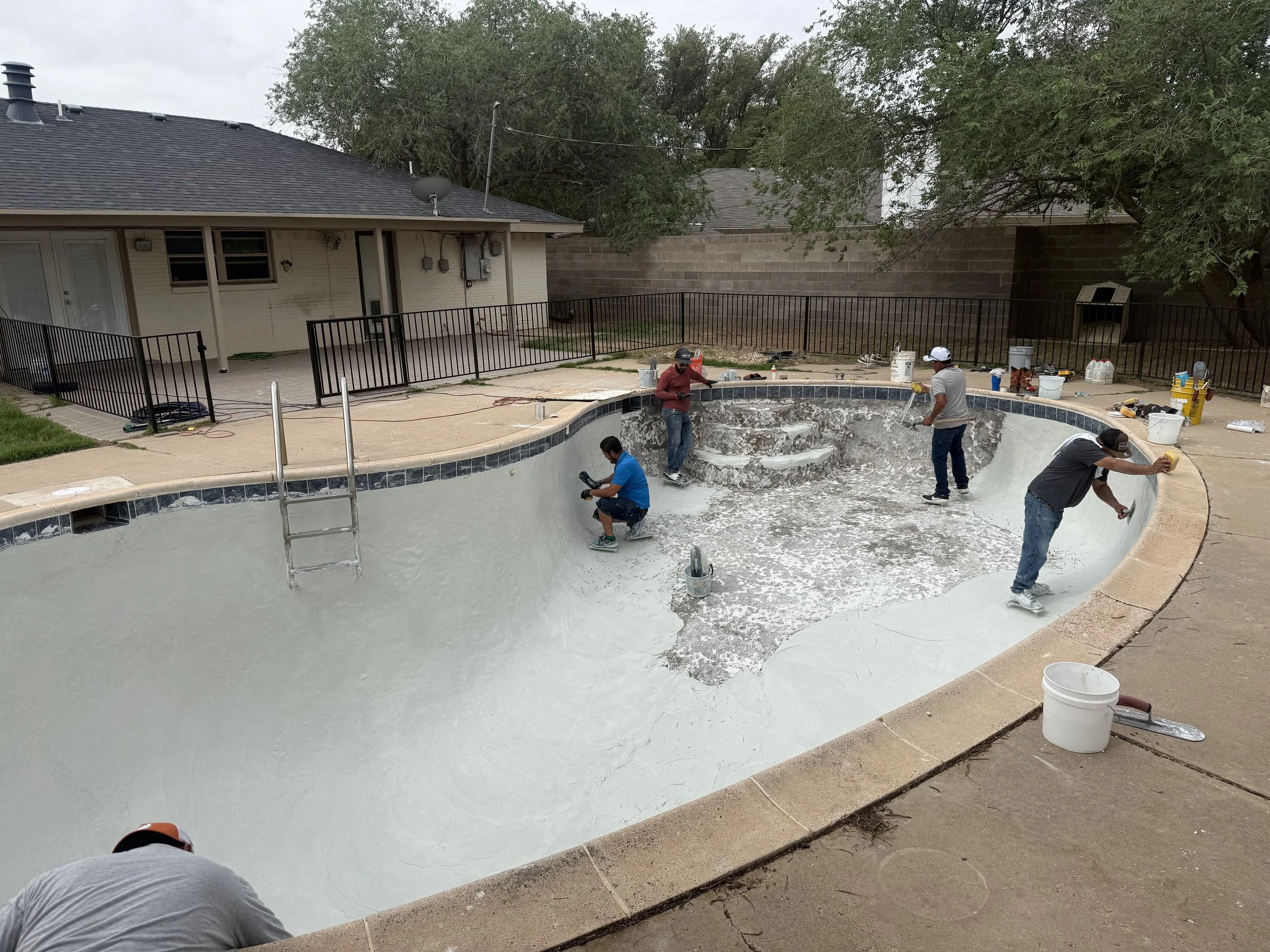 People working on resurfacing  a residential swimming pool in a backyard, with tools and supplies around.
