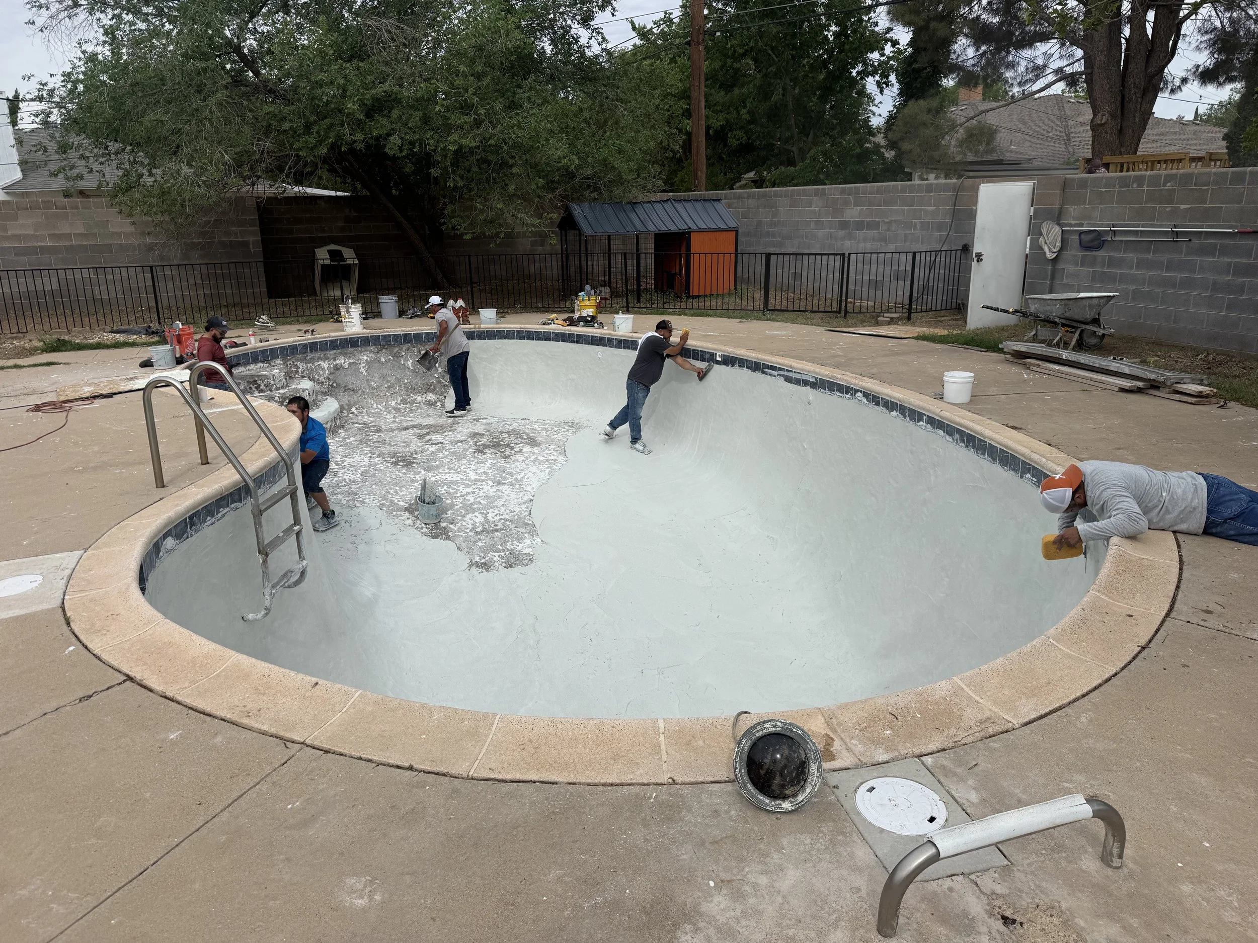 Workers refurbishing a backyard swimming pool by painting and preparing the surface. The empty pool has a curved shape with tiles lining the top edge. Several workers are applying paint, using tools, and working around the pool area, which is bordere