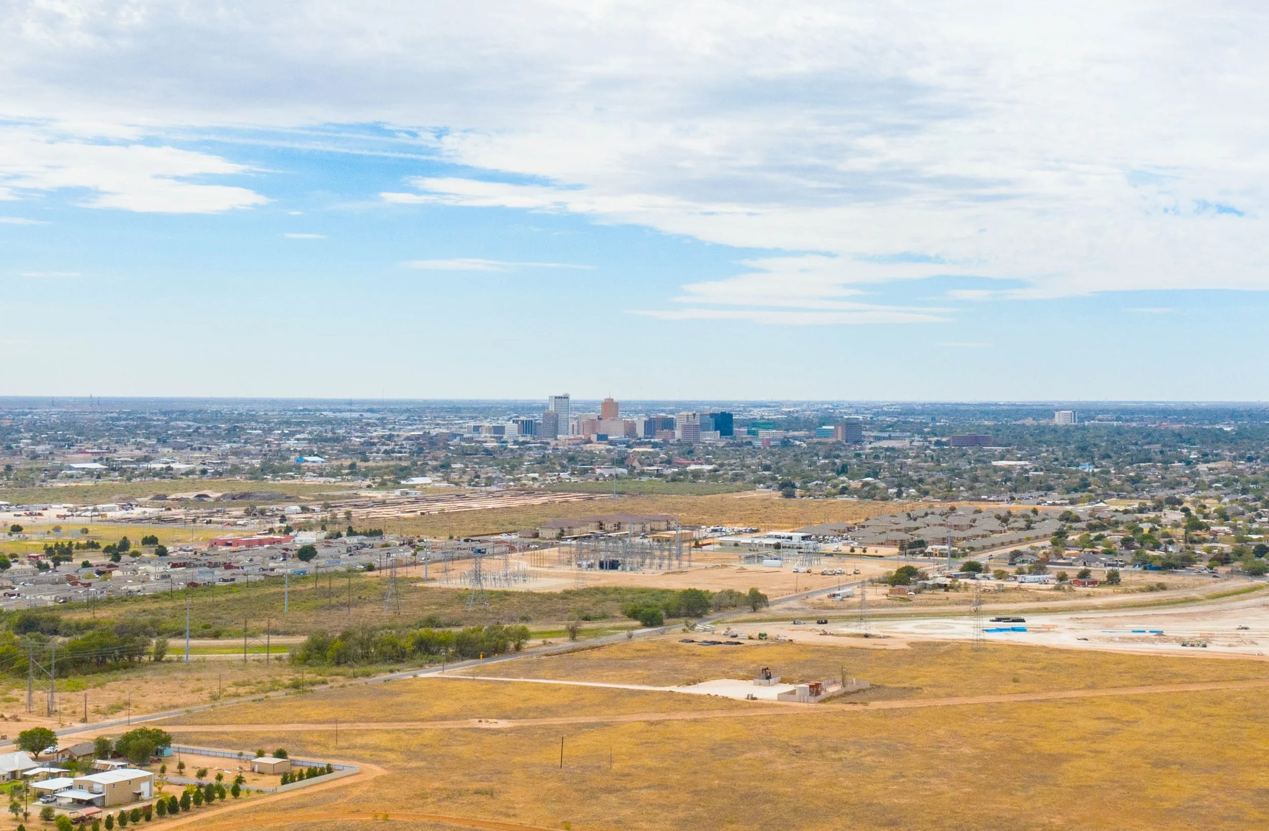 A panoramic view of a Midland, TX with tall buildings in the distance, under partly cloudy sky, with a foreground showing residential and commercial areas and open land.