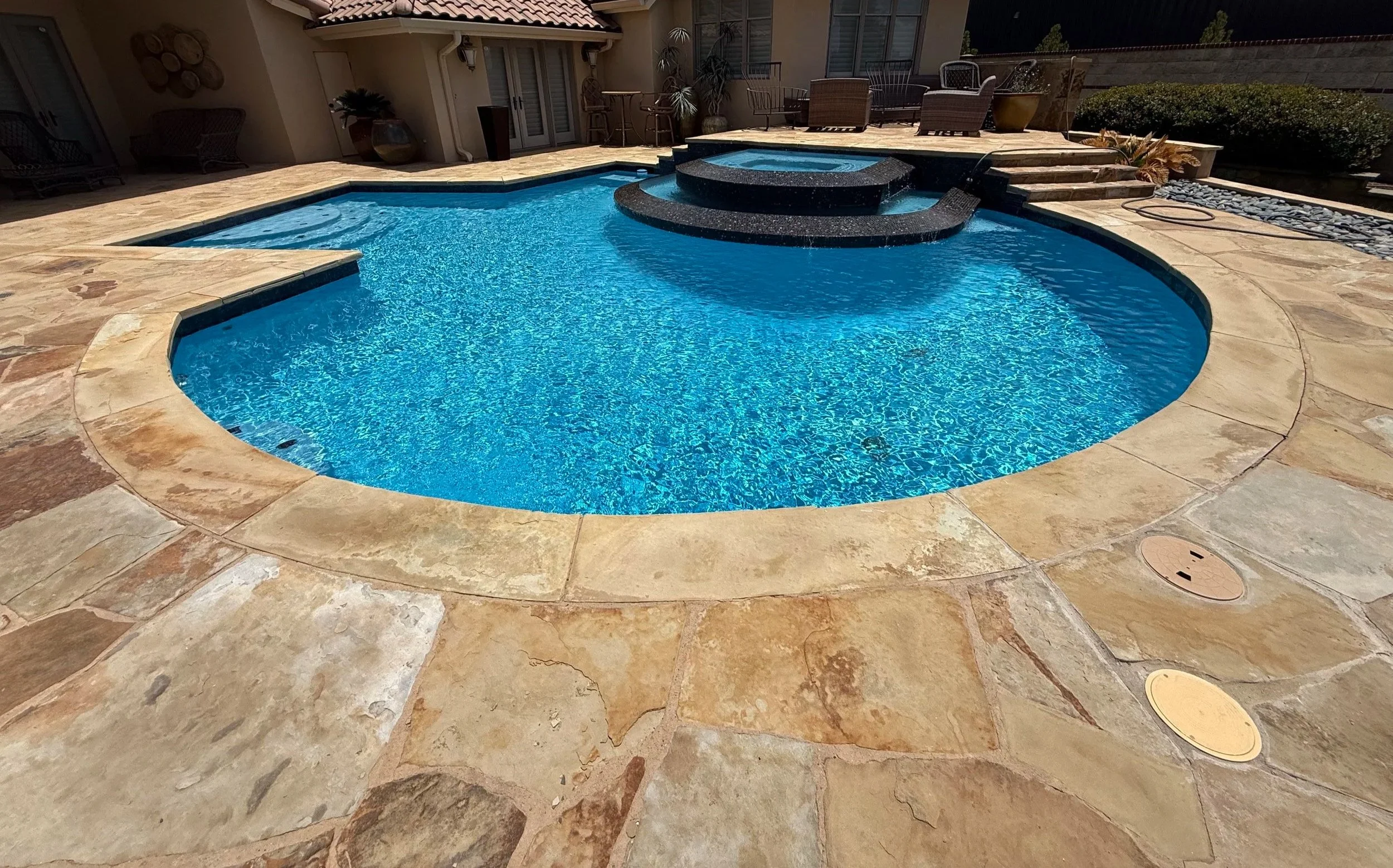 A backyard with a swimming pool with a spa, surrounded by a stone deck, outdoor furniture, and potted plants, with a house in the background.