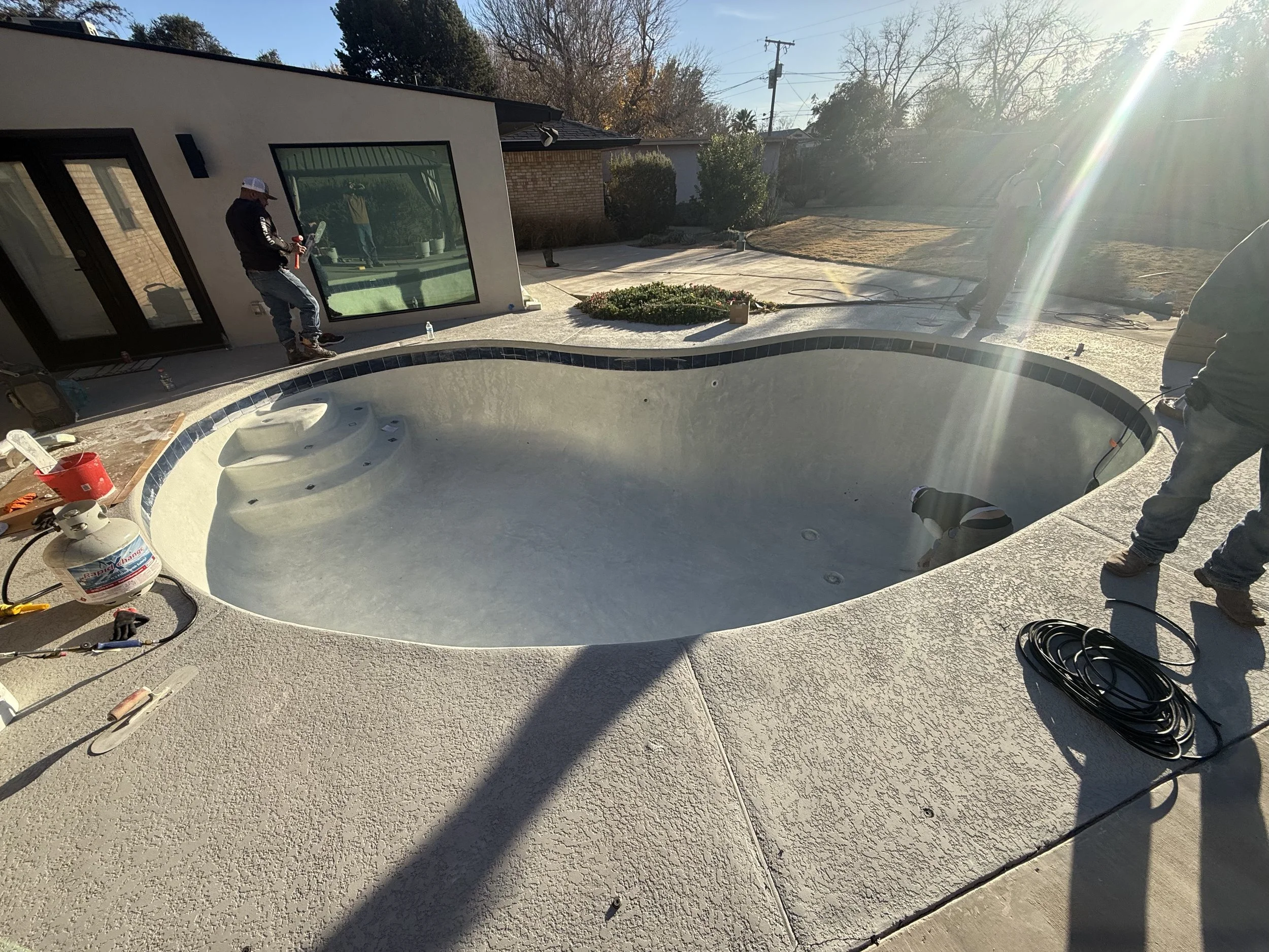 A backyard with a swimming pool, some construction tools, and workers installing a large window on the house. The sun is shining brightly, creating lens flare.