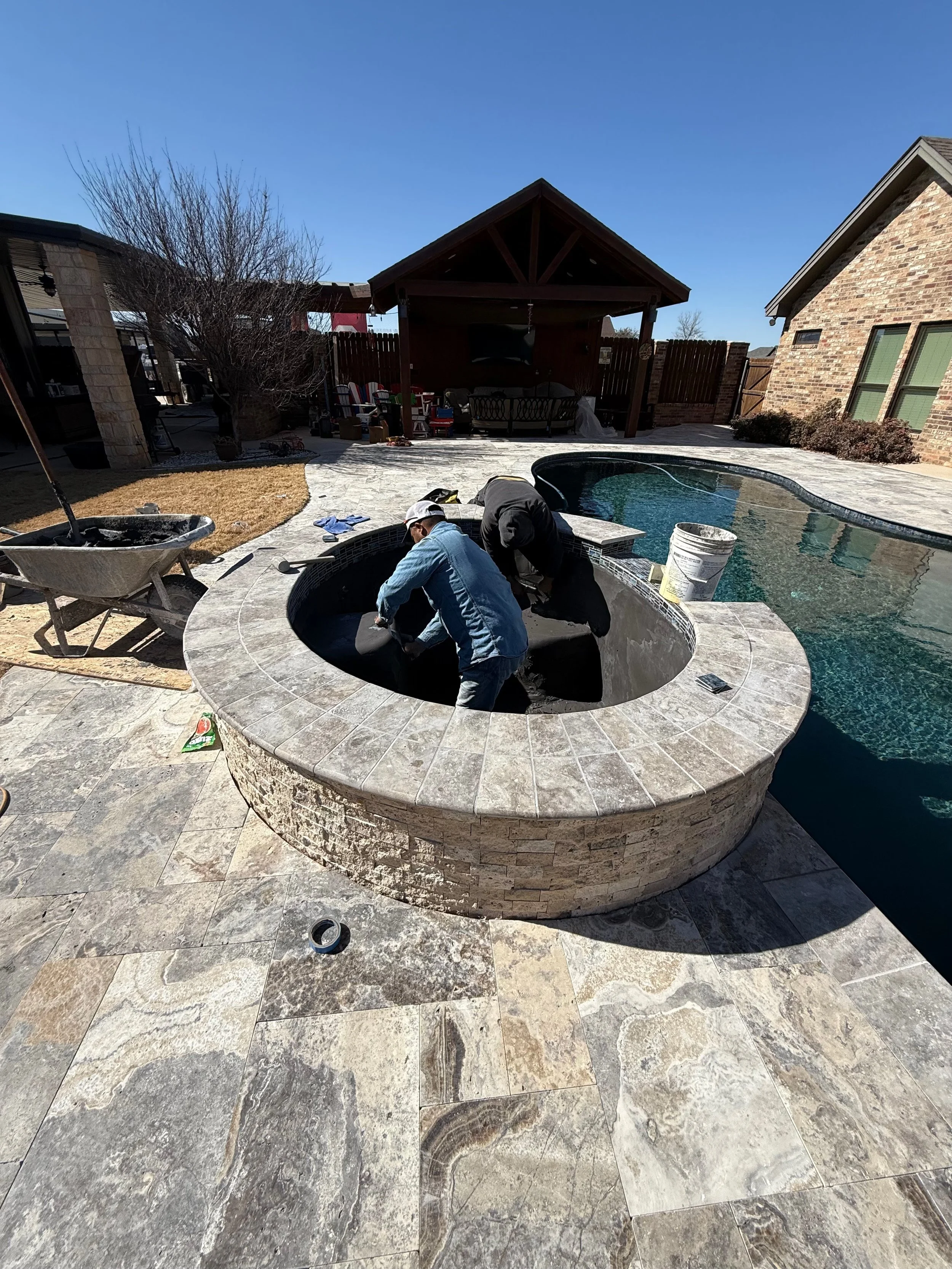 People working on a pool, hot tub, and stone patio, under a pergola, with outdoor furniture and tools.