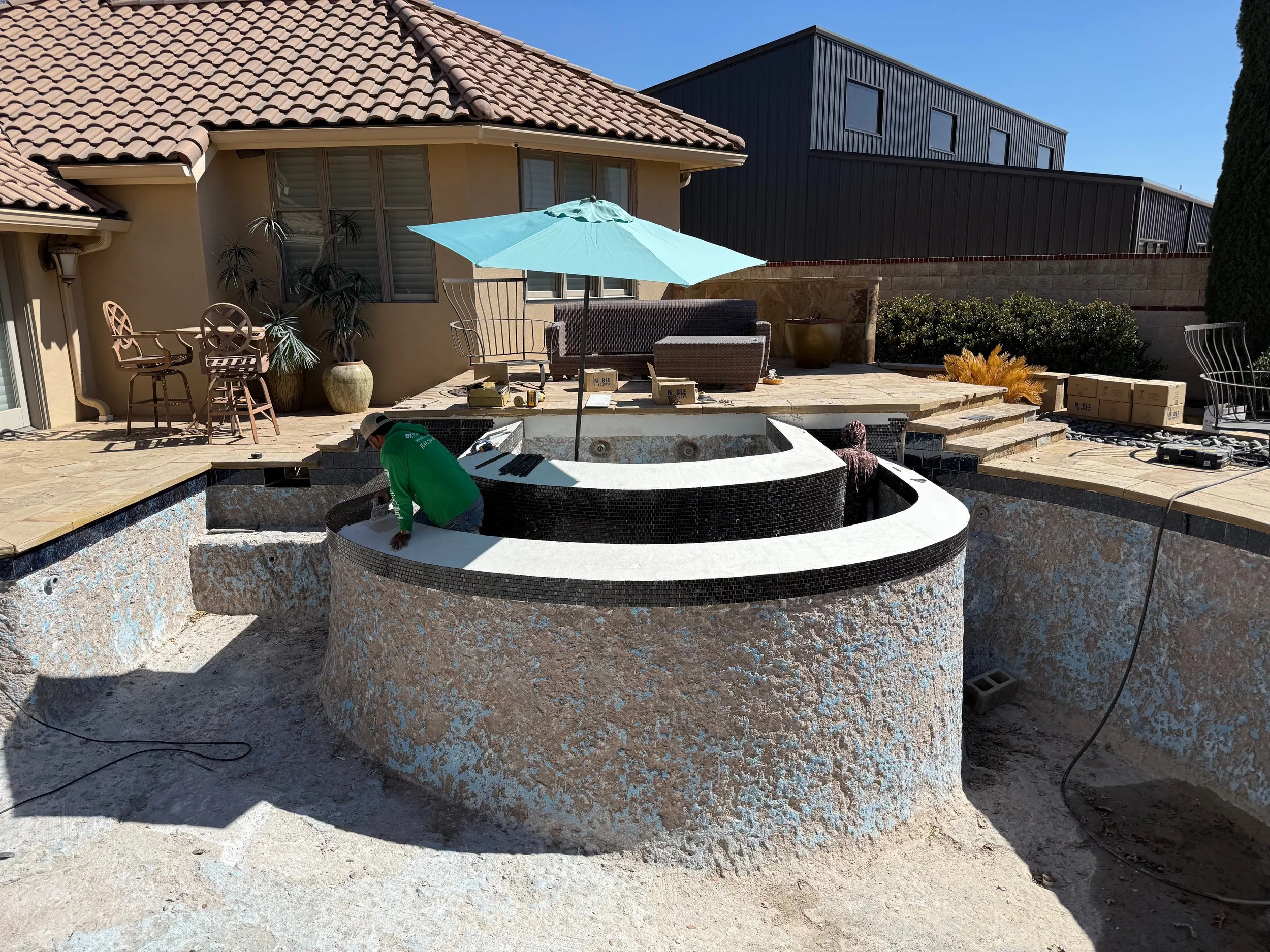 Backyard patio with pool under construction, with seating area, an umbrella, and construction tools, during sunny weather.