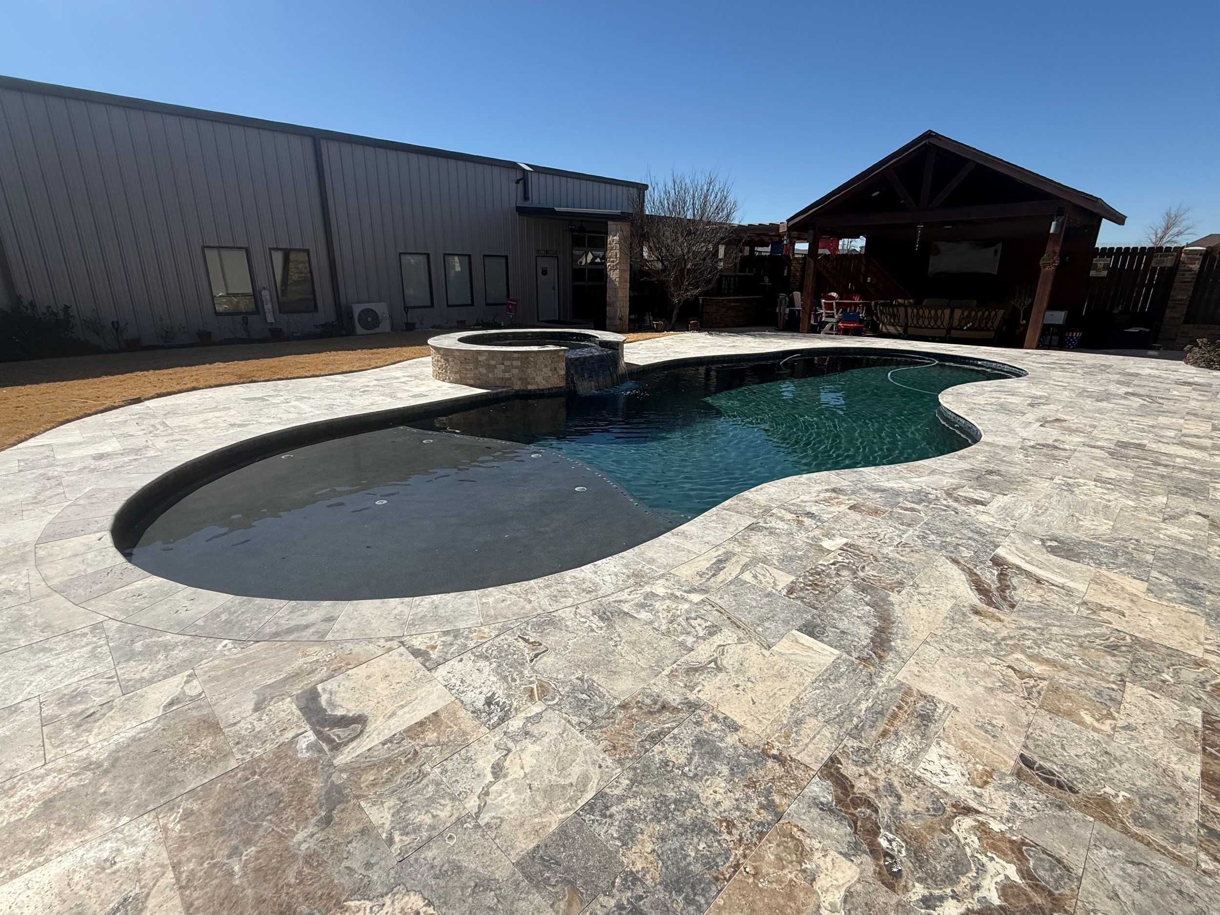 A backyard with a swimming pool, stone paving, a wooden gazebo with furniture, and a building in the background under a clear blue sky.