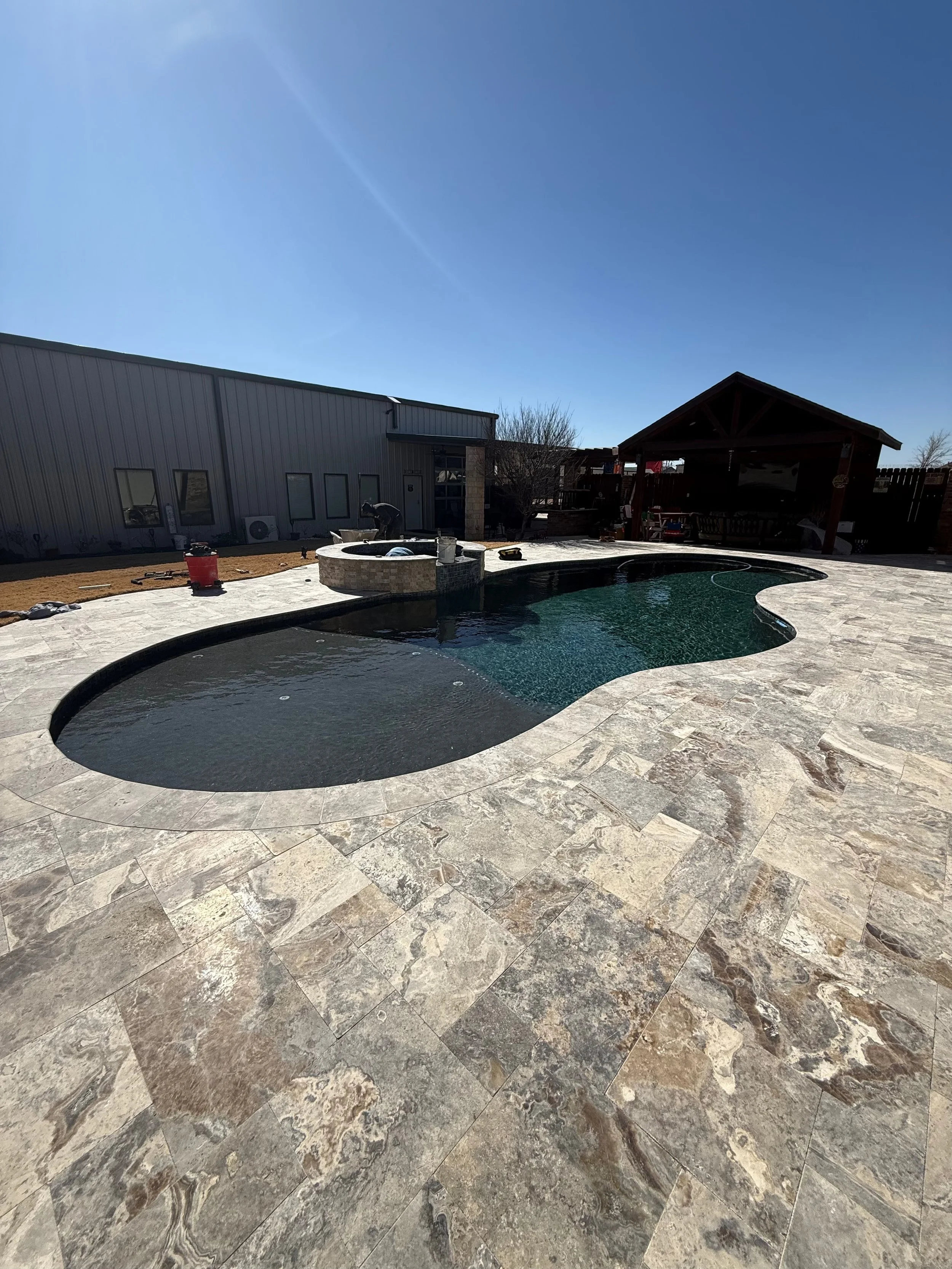 Backyard with a swimming pool, stone patio, a small pavilion with a bench, and a building nearby under a clear blue sky.