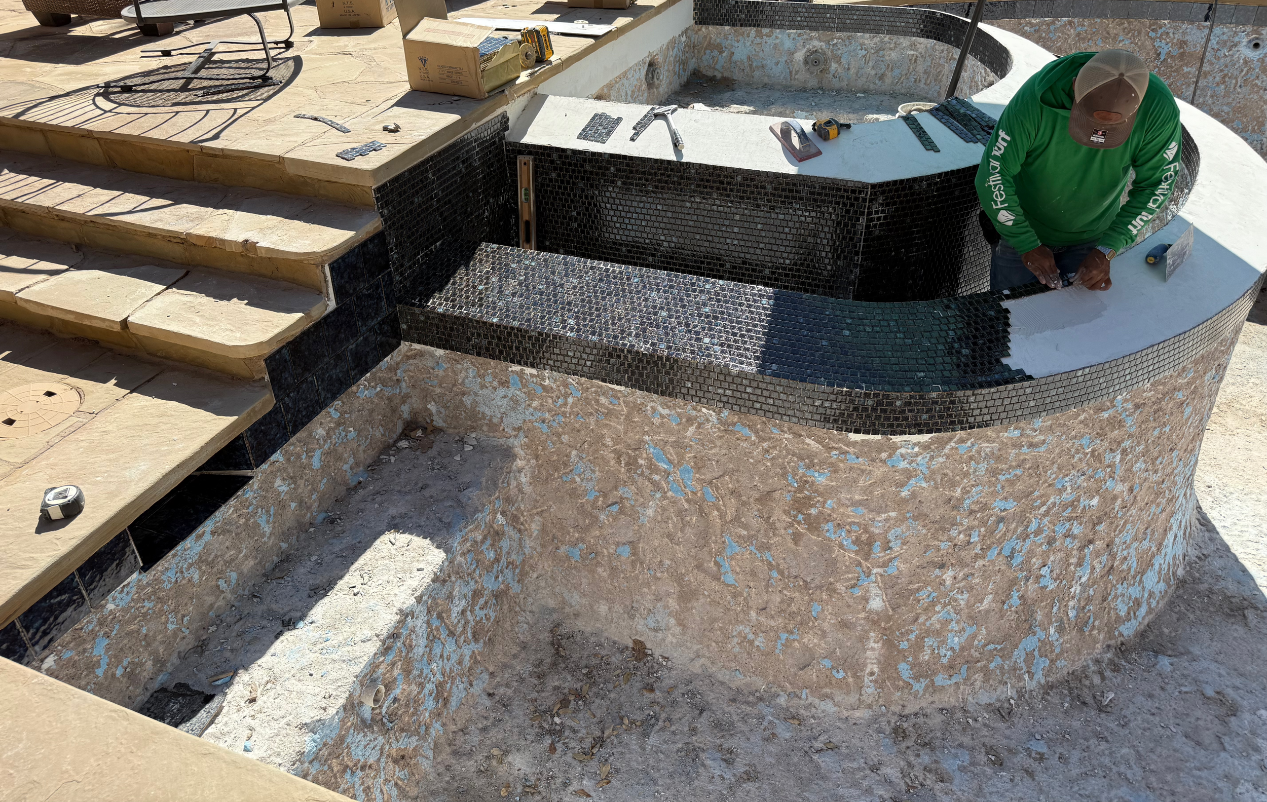 A person installing black mosaic tiles around an outdoor hot tub with beige steps nearby, tools and a box of tiles on the edge of the hot tub.