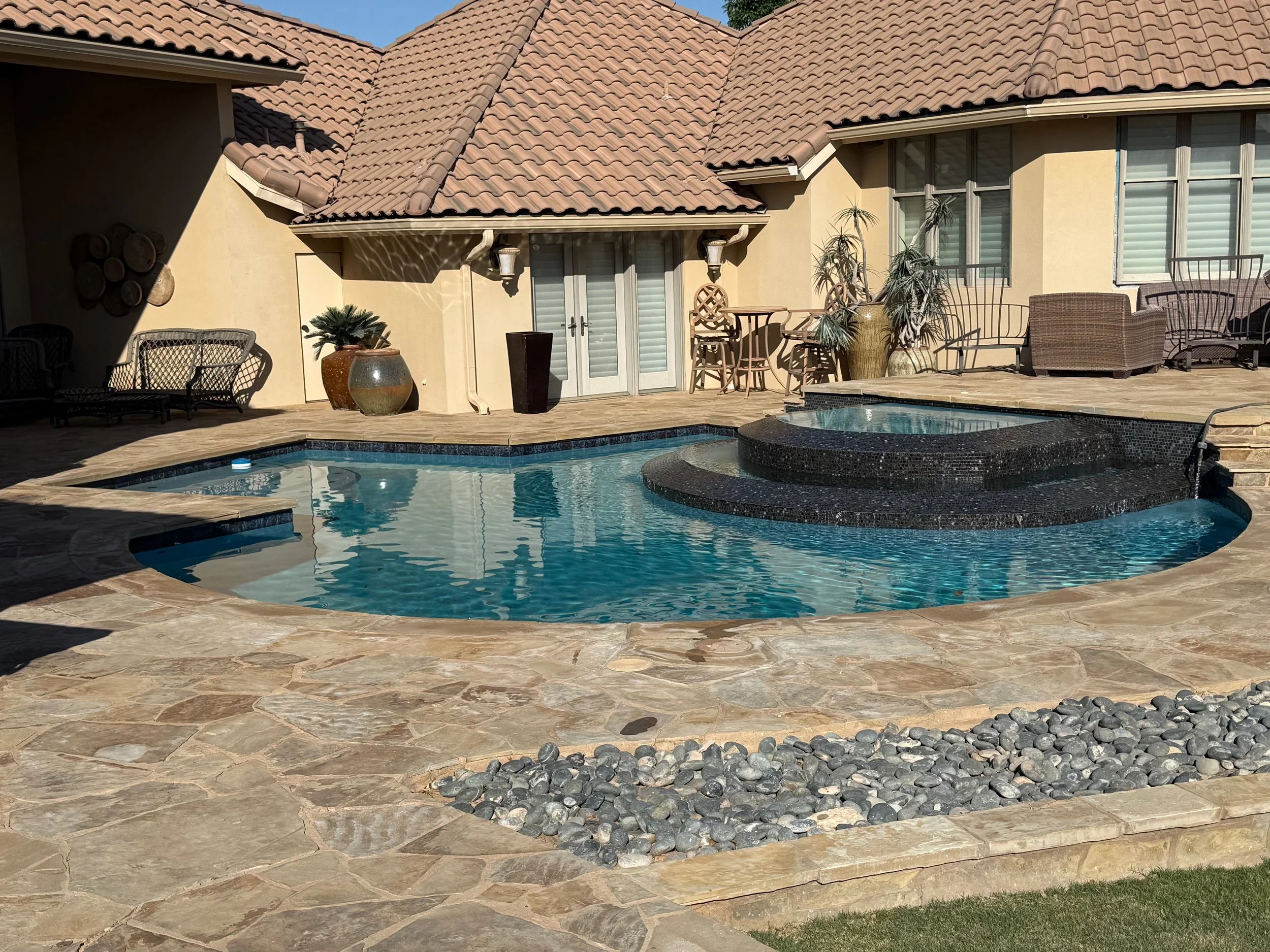 A backyard scene featuring a swimming pool with a raised hot tub, patio furniture including chairs and a table, large potted plants, and a house with a tiled roof and sliding glass doors.