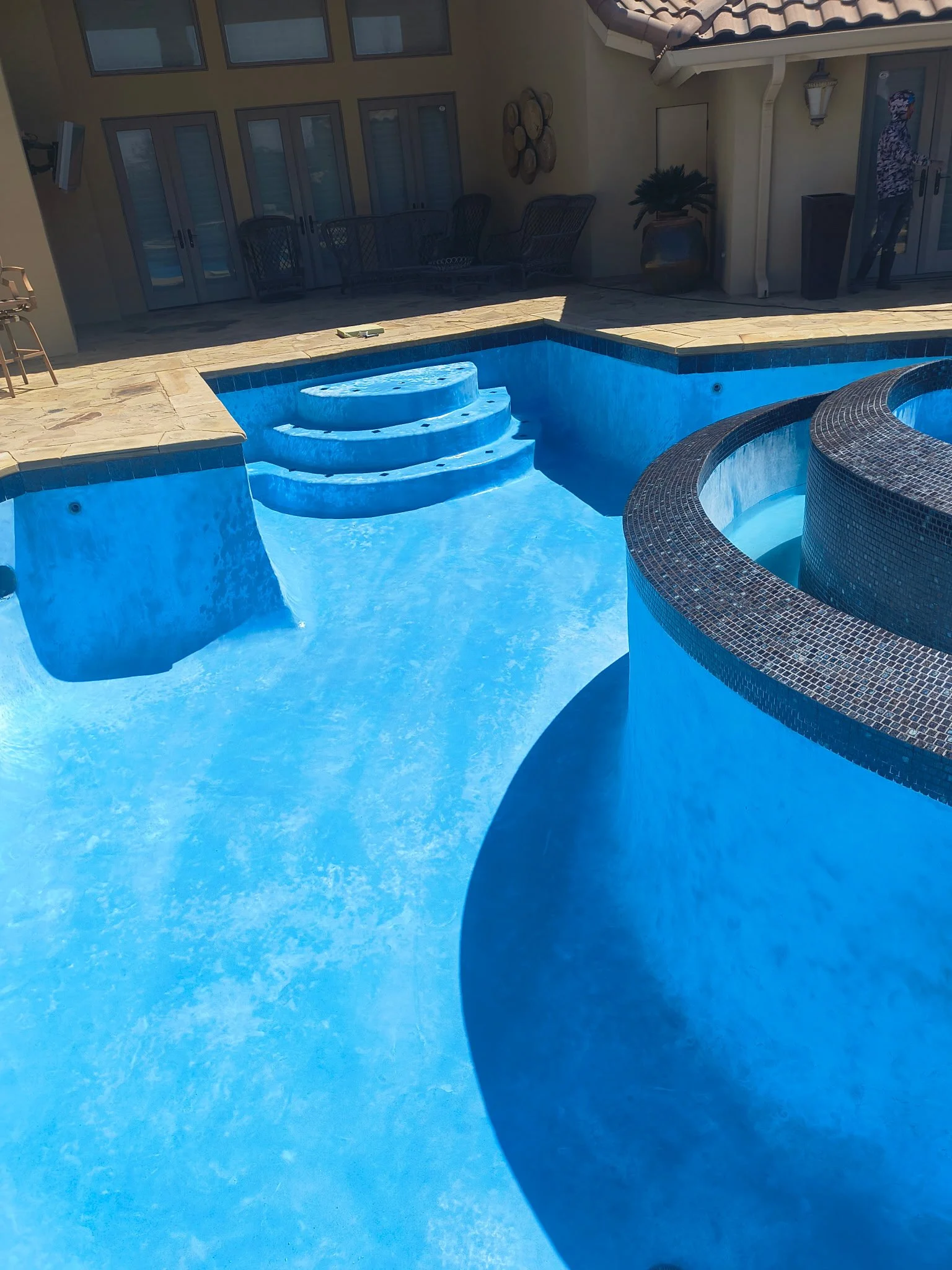 Backyard swimming pool with blue water, steps leading into the pool, and a hot tub with dark tile tiling. Lounge chairs, patio furniture, a potted plant, and a person near the sliding glass doors are visible in the background.