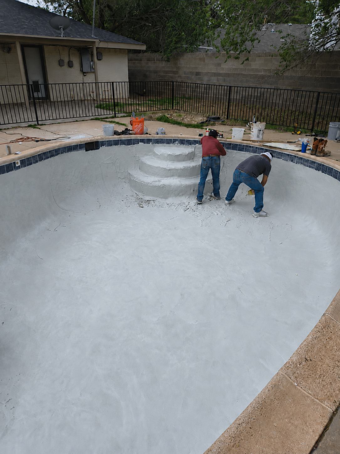 Two workers are refinishing the interior of an empty swimming pool, applying a white coating or plaster.