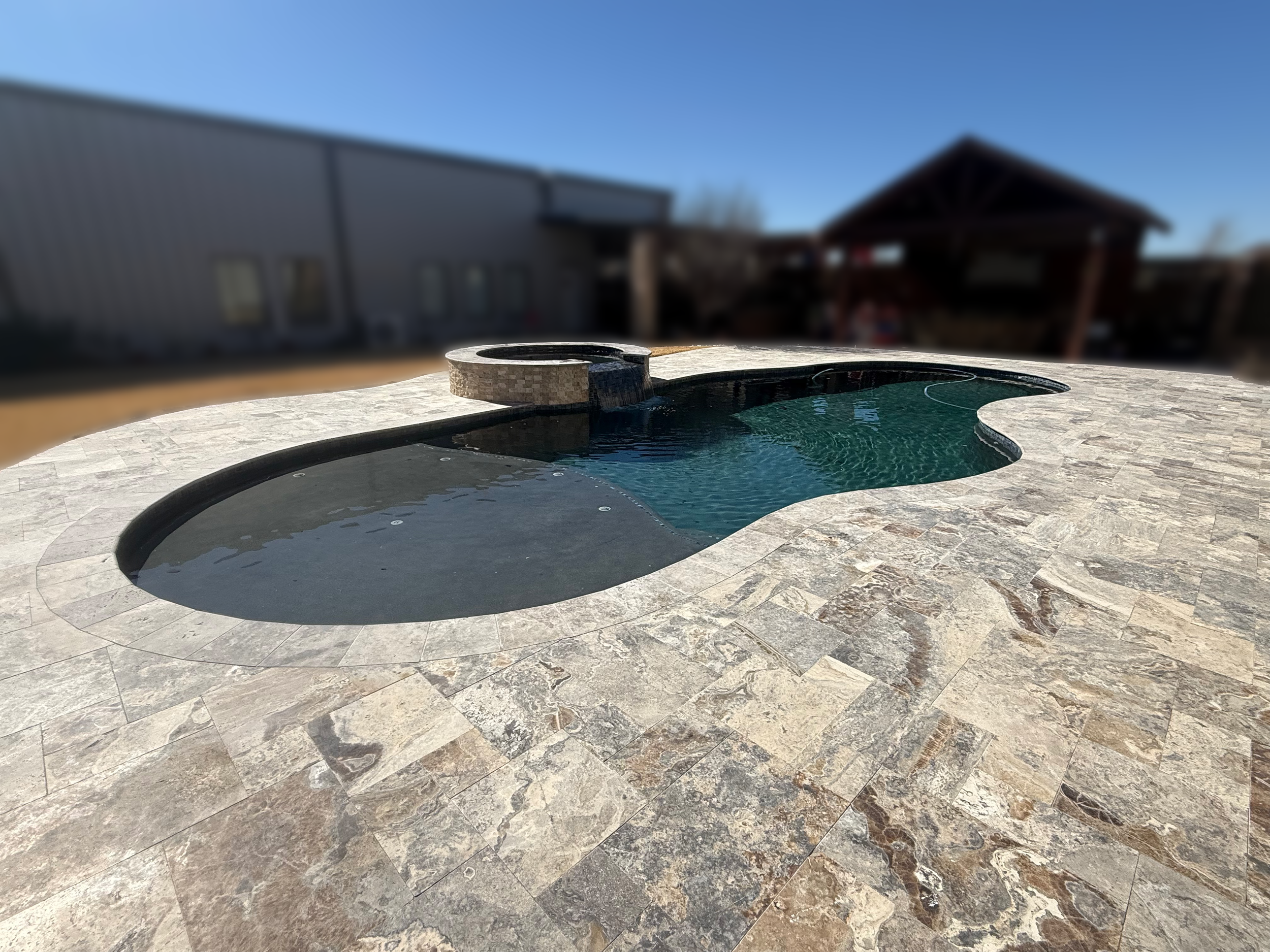 A backyard with a swimming pool, surrounded by stone patio and a building with a tan exterior and large windows, under a clear blue sky.