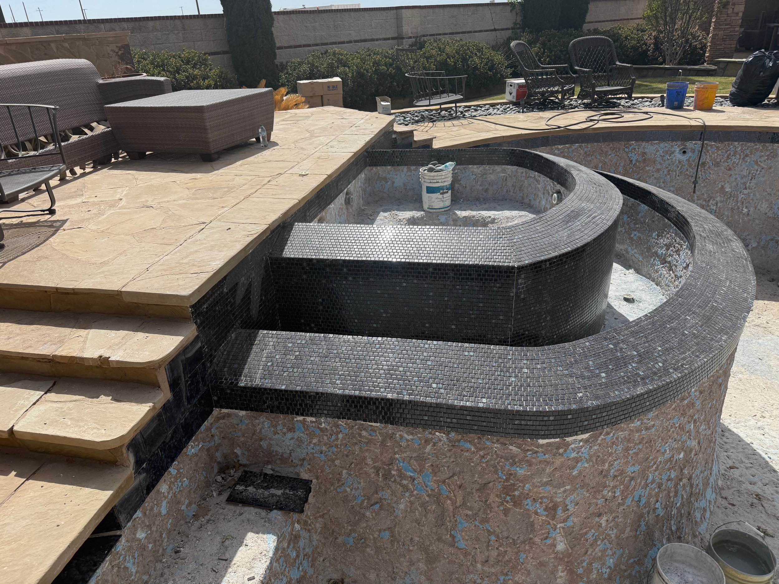 This image shows a backyard patio area under construction. There are outdoor furniture pieces, including a trash can, black chairs, buckets, and a coiled hose. The hot tub or pool area appears to be in the process of renovation, with some tiles missi