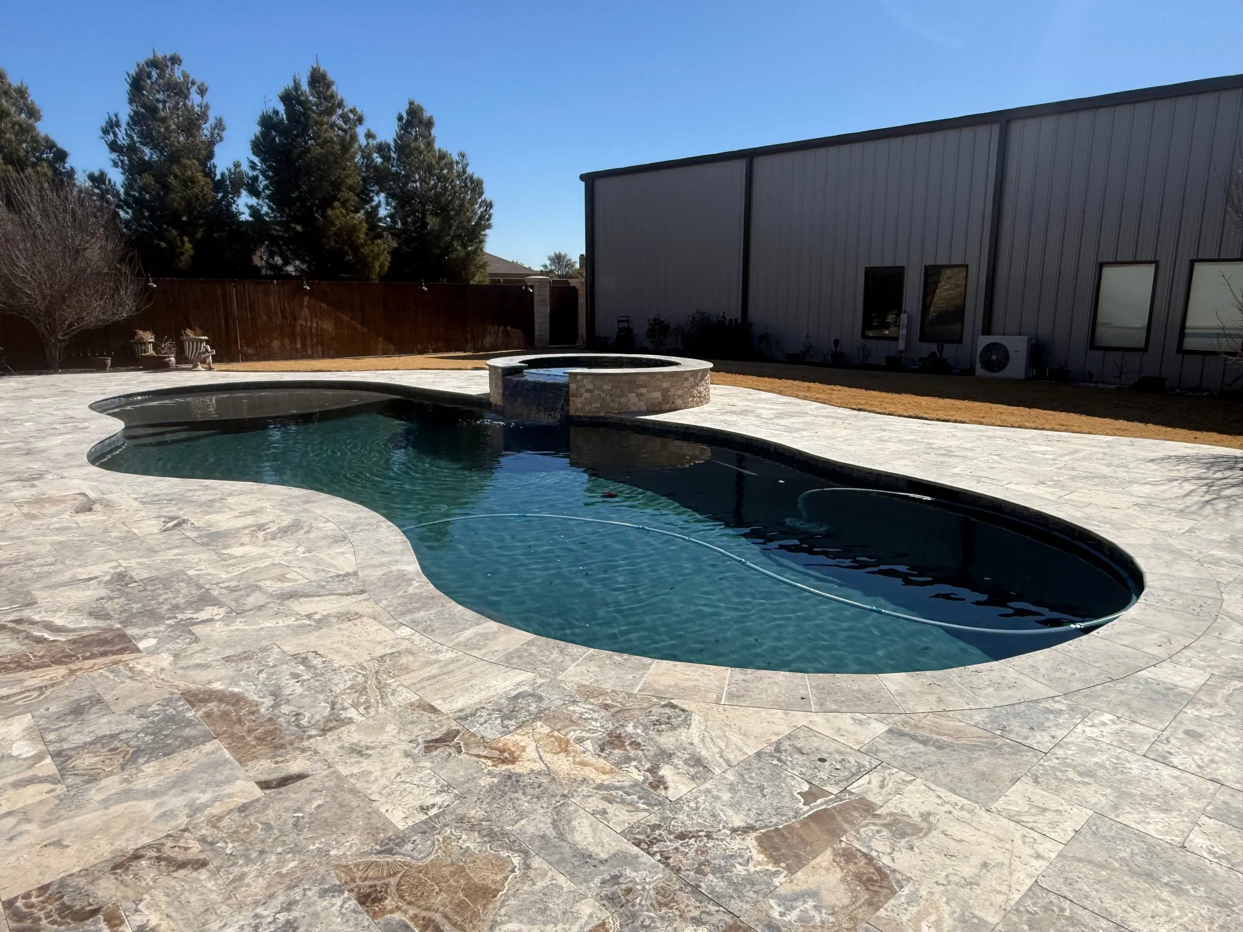 A backyard with a kidney-shaped swimming pool, a stone fire pit, a patio with stone tiles, a wooden fence, and some trees and bushes.