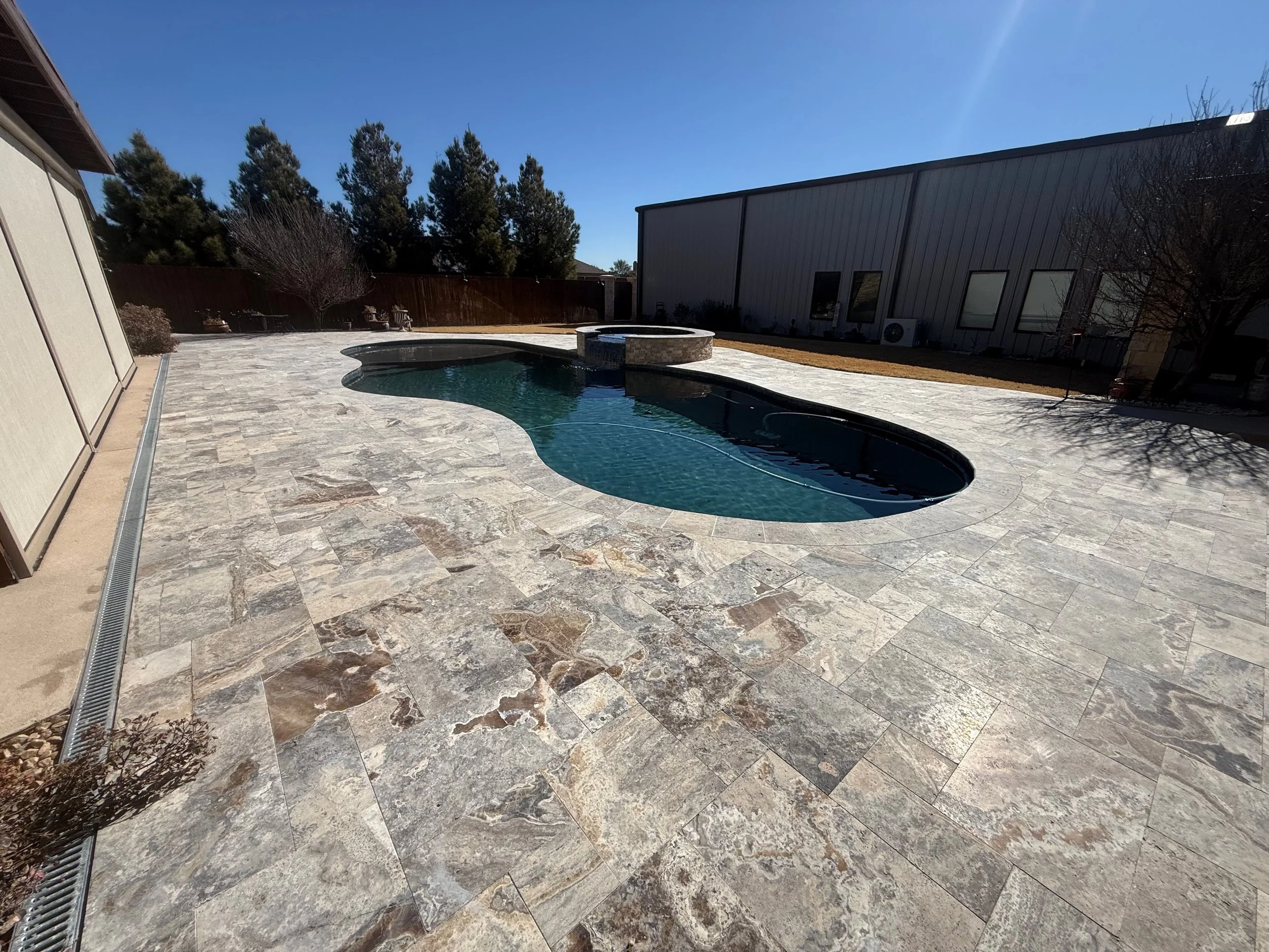 A backyard with a stone pool, a hot tub, and a paved patio area, surrounded by a wooden fence and trees under a clear blue sky.