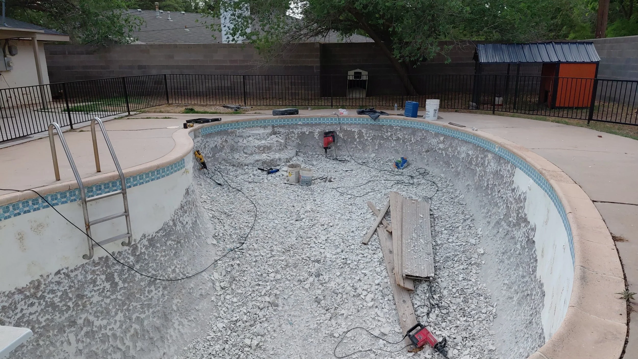 Empty swimming pool under construction with debris, tools, and buckets inside, surrounded by a concrete deck and black metal fence, with trees and houses in the background.