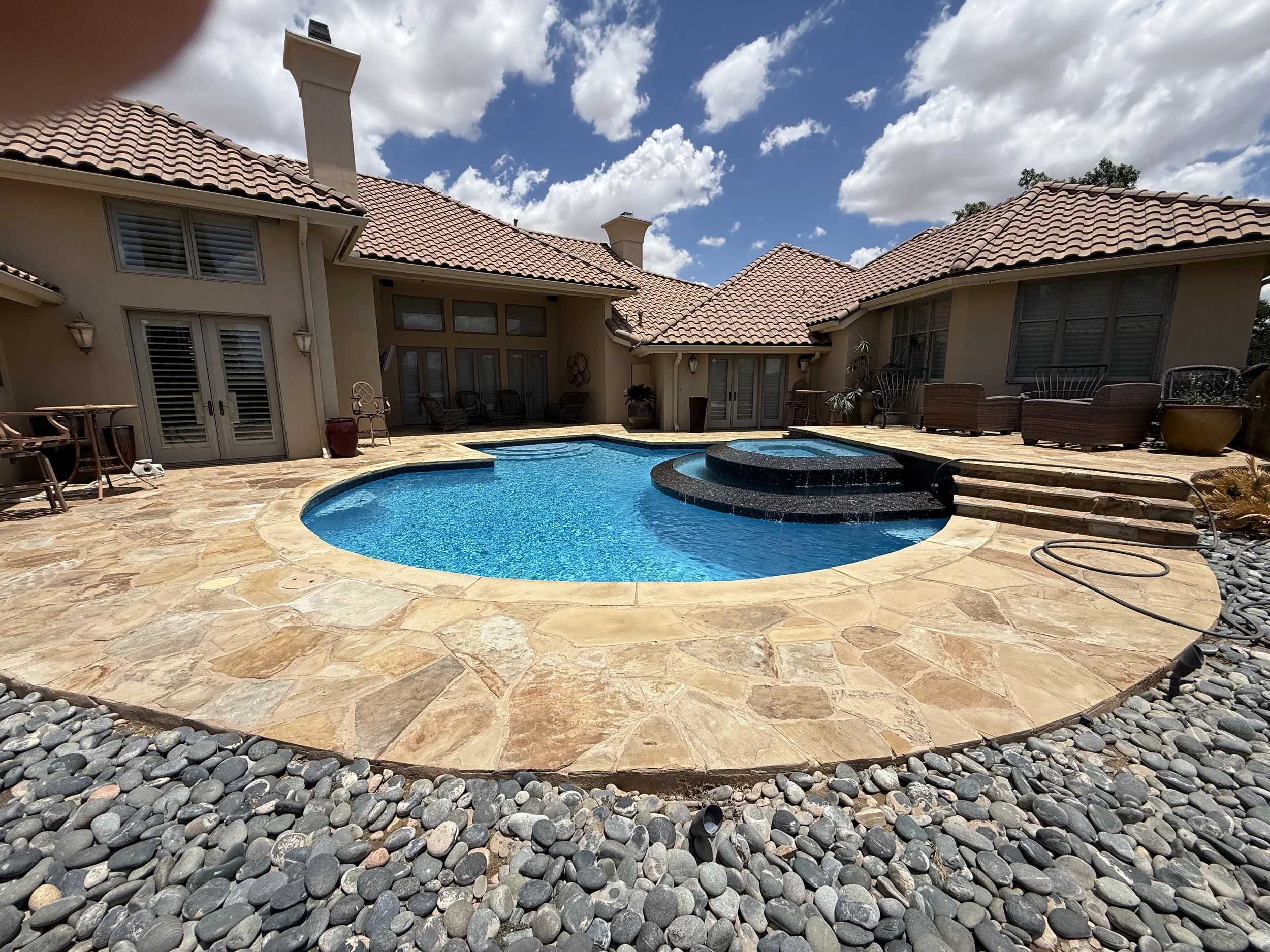 Backyard with a kidney-shaped swimming pool, surrounded by a stone patio and black river rocks, with seating and potted plants next to a house with red tile roof and beige exterior walls.