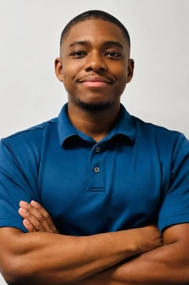 Young man with short hair and a goatee smiling, wearing a blue polo shirt with arms crossed against a plain white background.