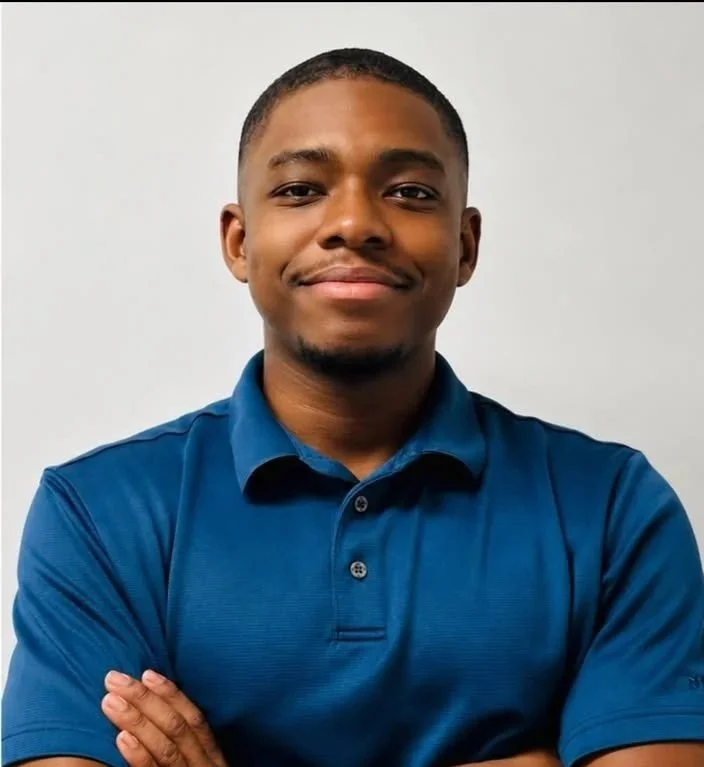 A young African American man with short black hair, wearing a blue polo shirt, smiling with arms crossed against a plain light gray background.