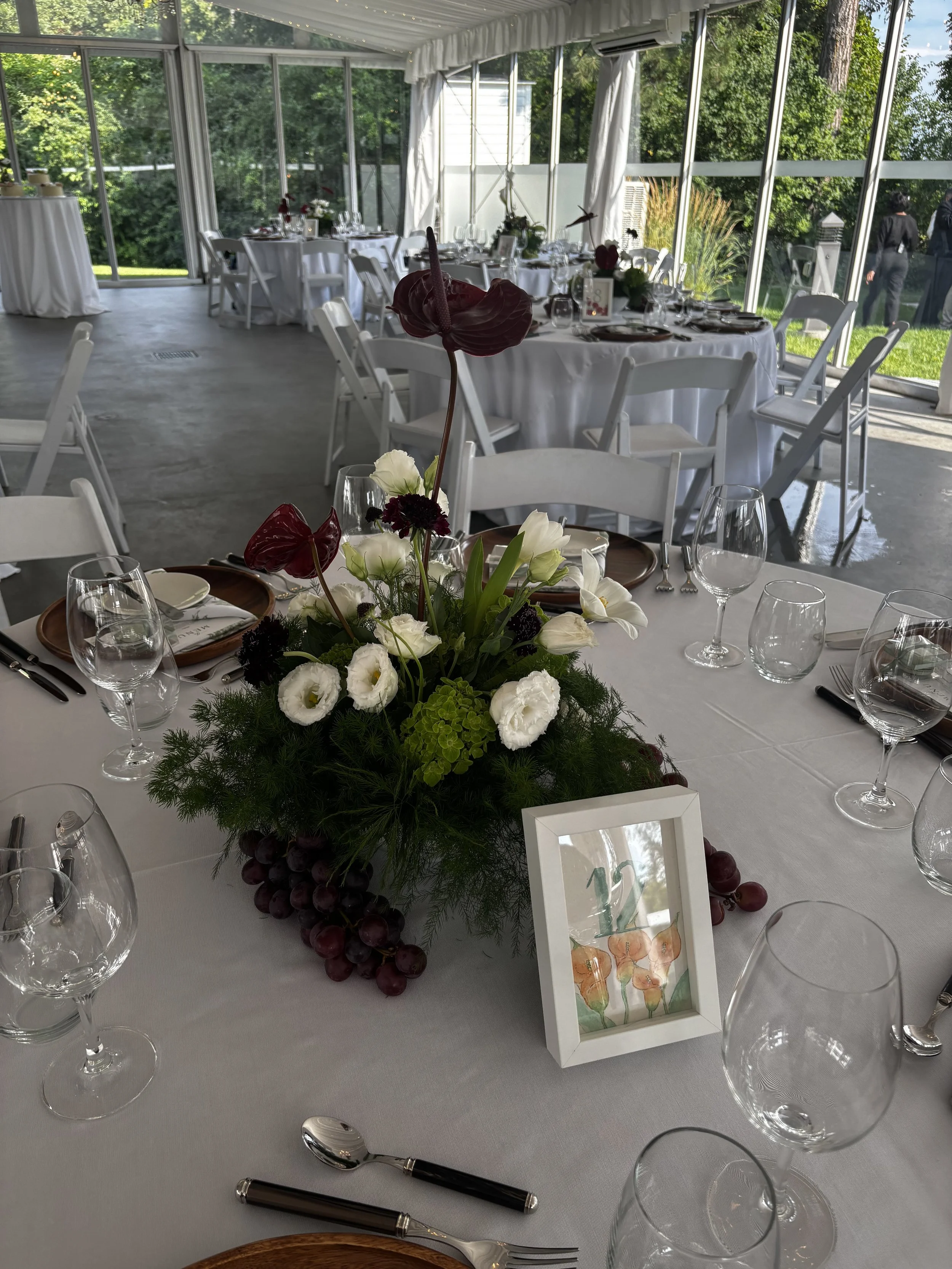 A table set for a formal event with a floral centerpiece, wine glasses, and tableware, inside a bright glass-enclosed venue with outdoor greenery visible through the windows.