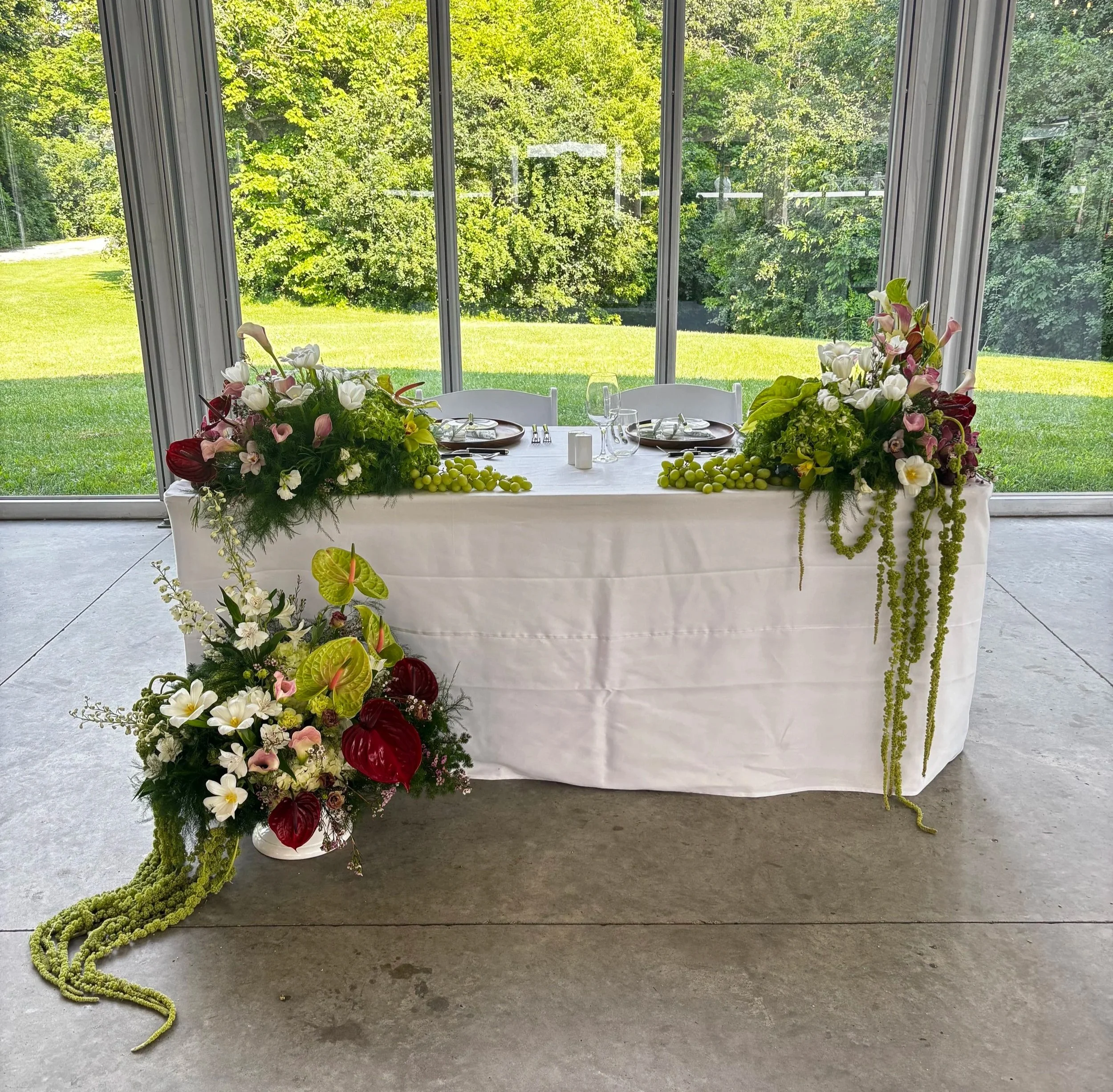 A decorated rectangular table with floral centerpieces and green grapes, set in front of large glass windows looking out onto a grassy yard with trees.