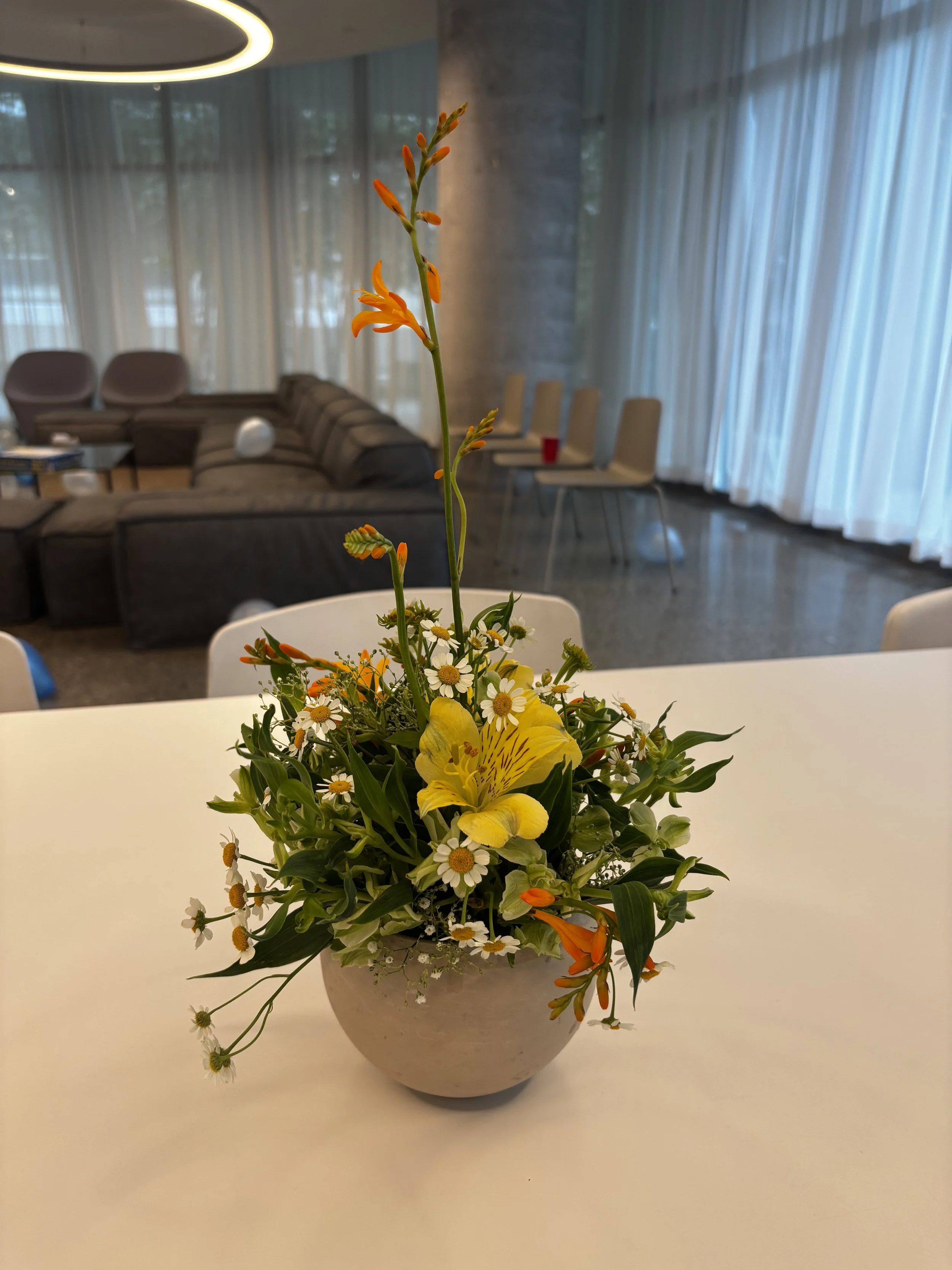 A floral arrangement featuring yellow lilies, white daisies, and small orange and yellow flowers in a white round vase, placed on a white table.