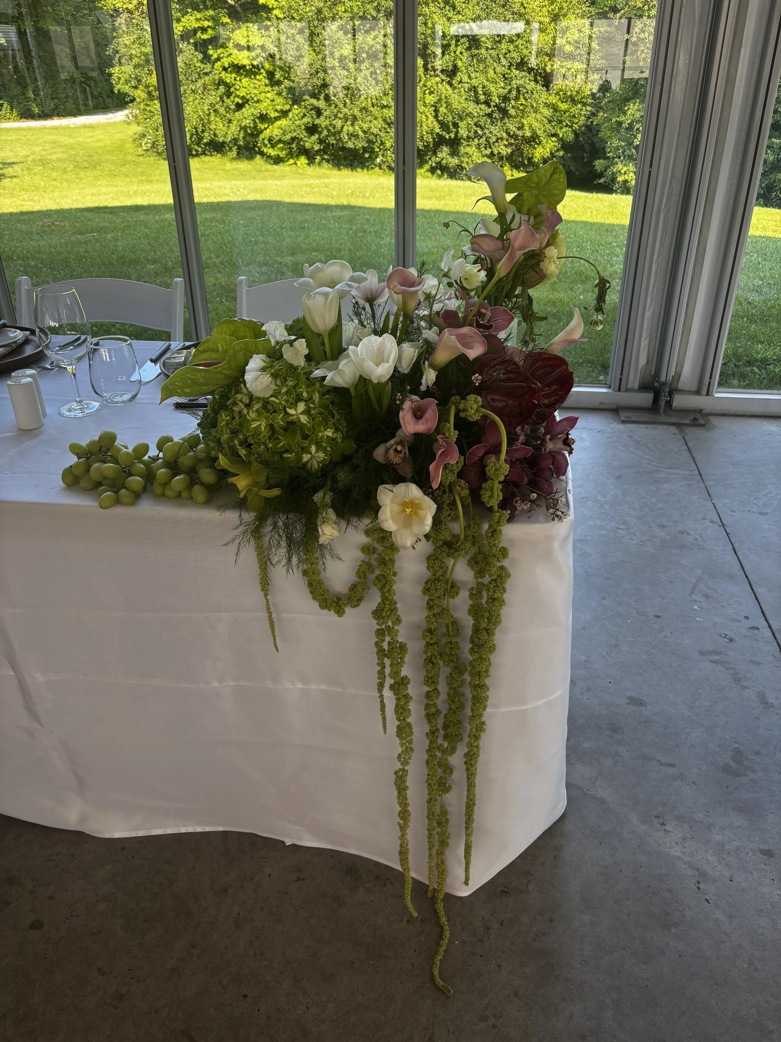 A white tablecloth with a floral centerpiece of white, pink, and purple flowers, with some green grape clusters and green hanging vines, set against large windows showing a green lawn and trees outside.