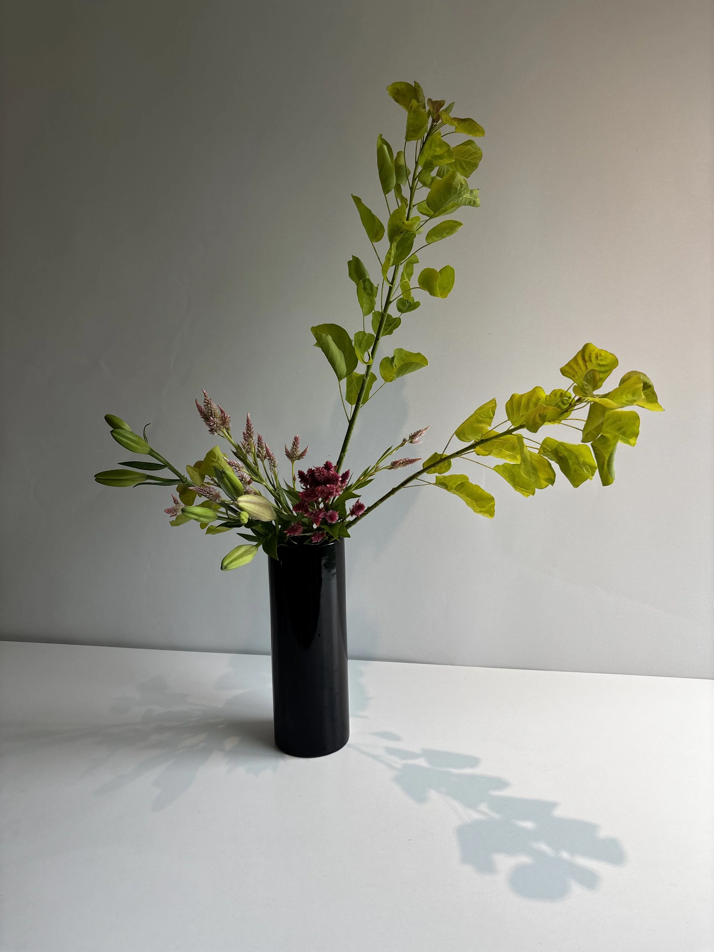 A black vase holding a colorful bouquet of flowers and green foliage against a plain white background.