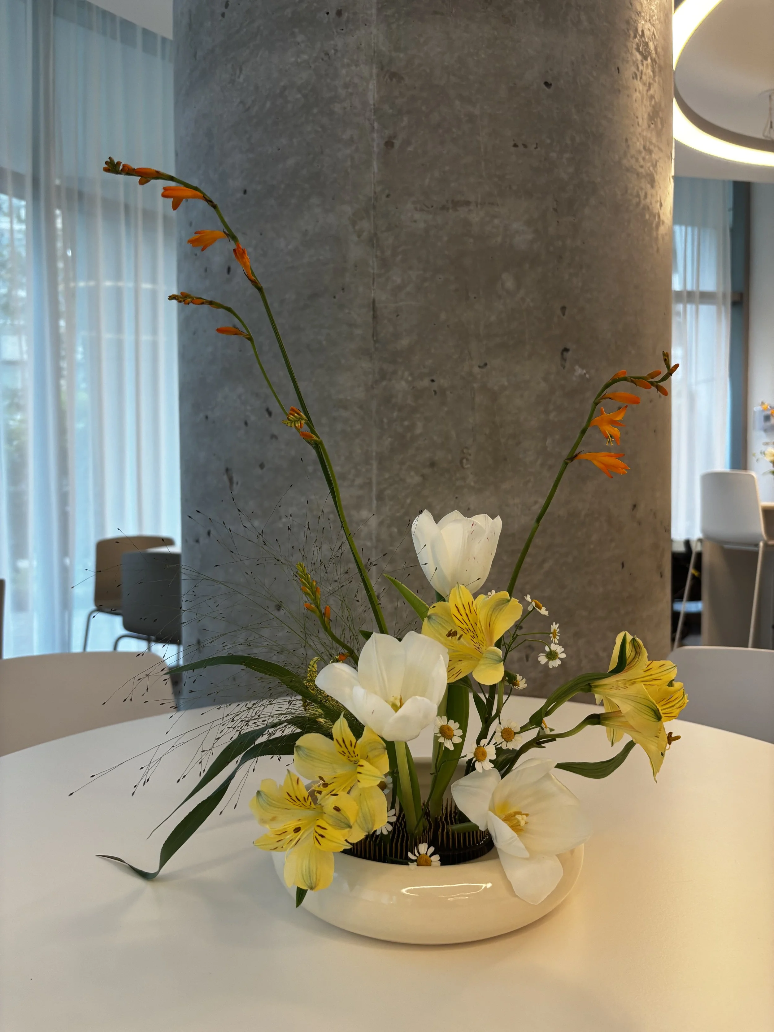 A floral arrangement with white, yellow, and orange flowers in a black vase on a white table, with modern interior and natural light in the background.