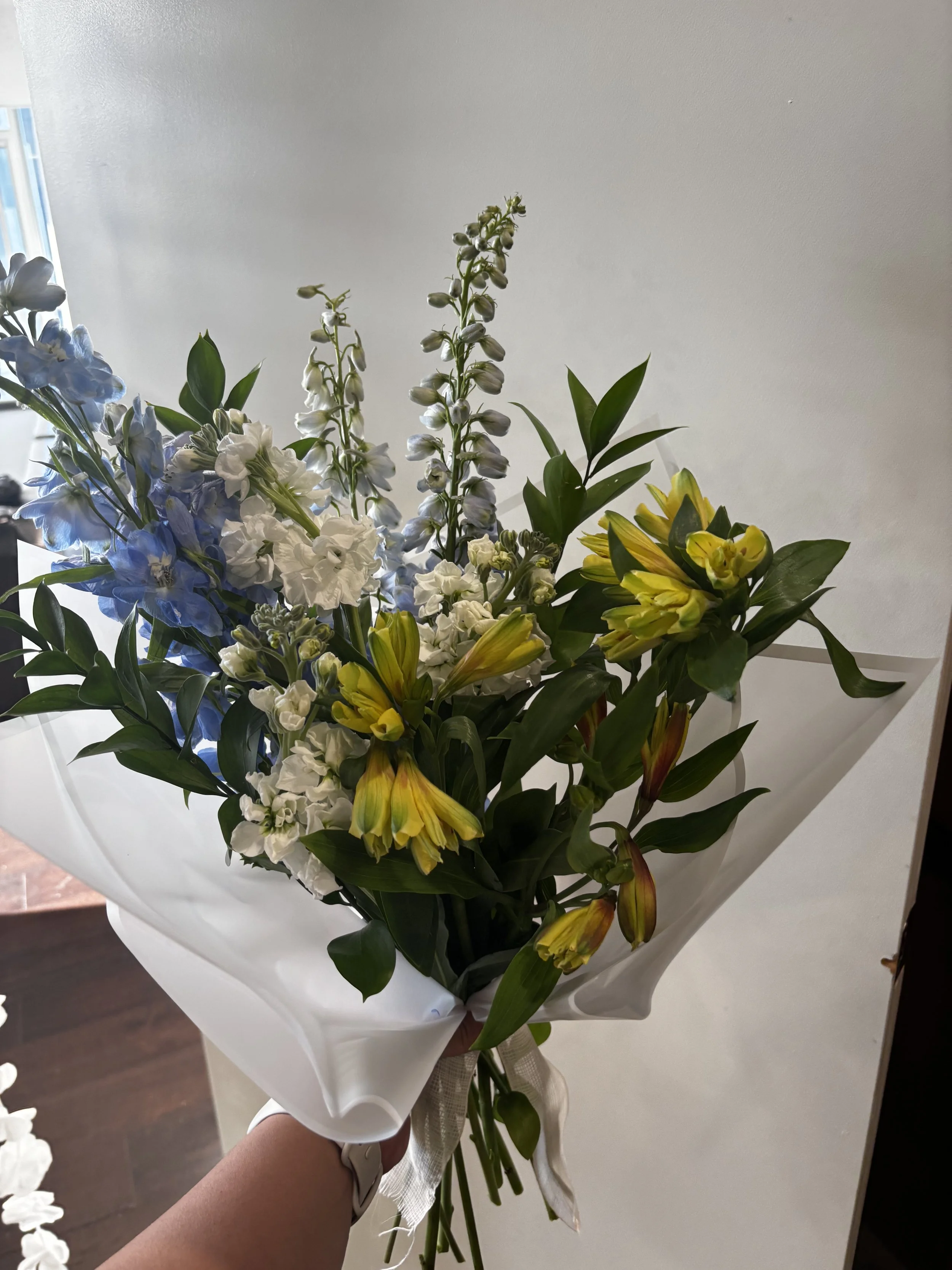 A hand holding a bouquet of mixed flowers with white, yellow, and blue blossoms, wrapped in white paper.