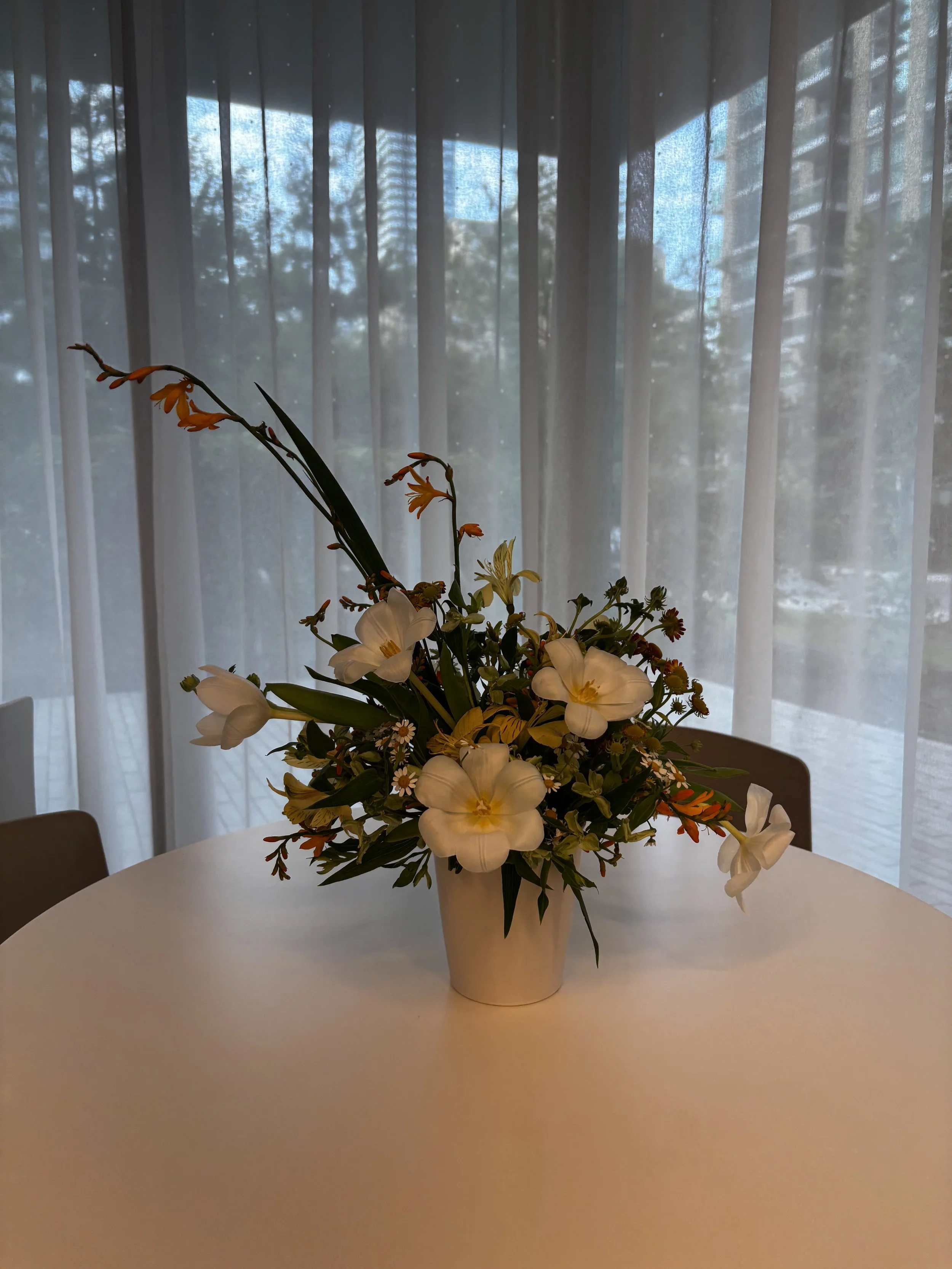 A bouquet of white and orange flowers in a beige vase on a white table, with a sheer curtain and trees visible outside in the background.
