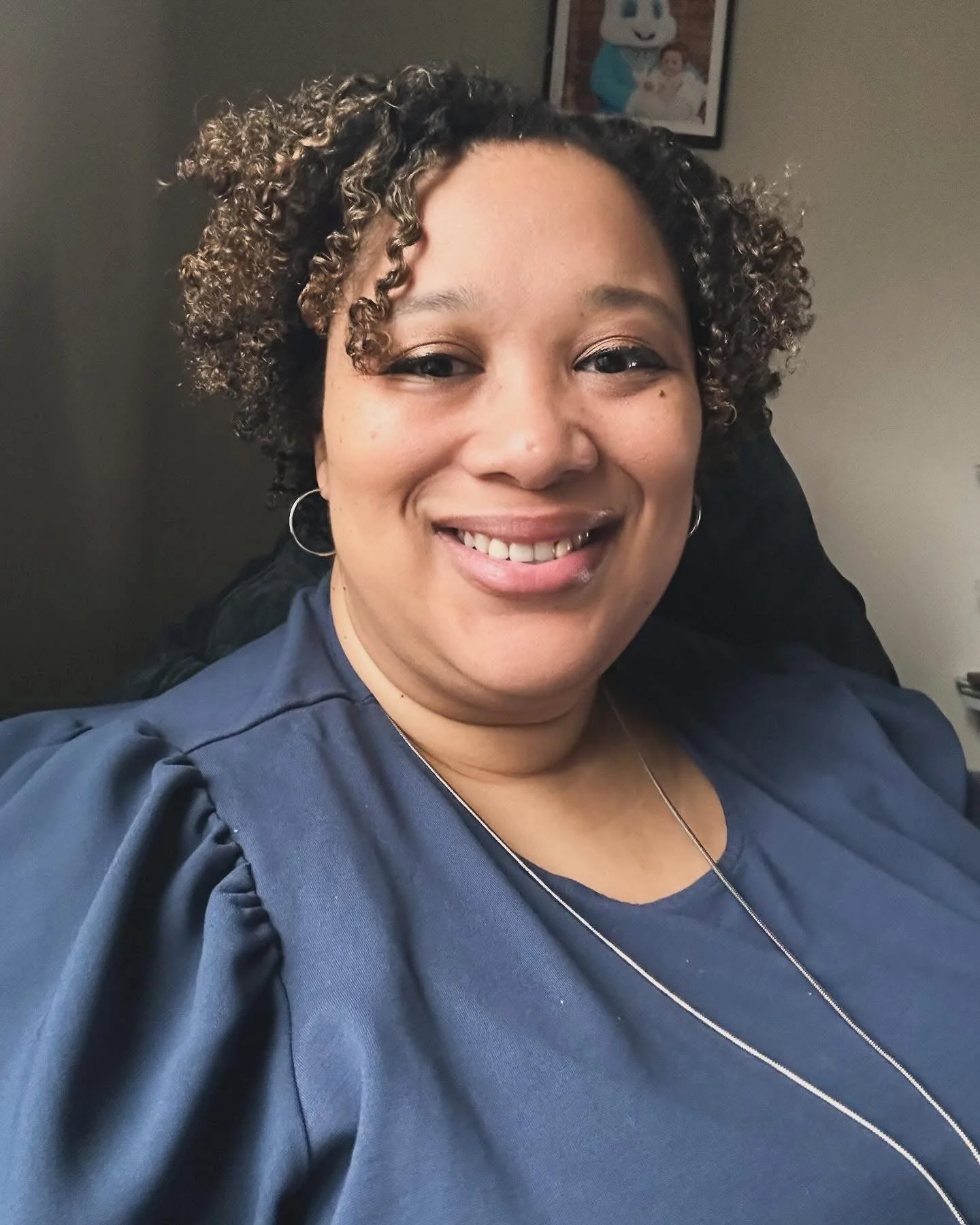 A smiling woman with curly hair, wearing earrings and a navy blue top with ruffled sleeves, sitting in an indoor setting with framed pictures on the wall behind her.