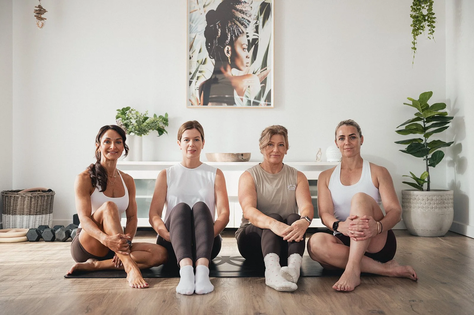 Four women sitting on yoga mats in a bright room with plants and artwork, ready for a yoga session.