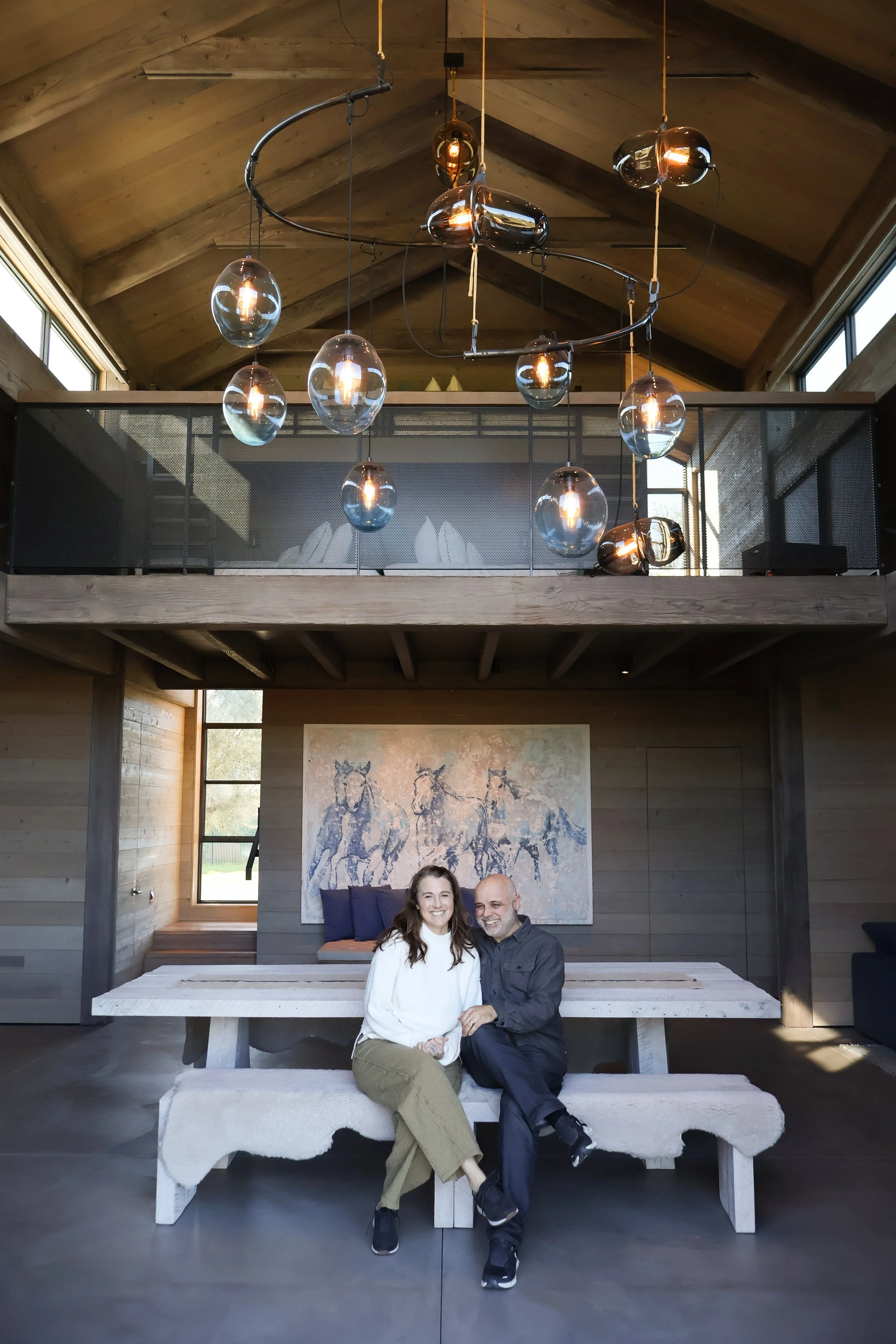 A man and a woman smiling and sitting on a large white bench with a rustic wooden wall and a large painting of horses behind them in a modern, wood-paneled room with a high ceiling and unique lighting fixtures.