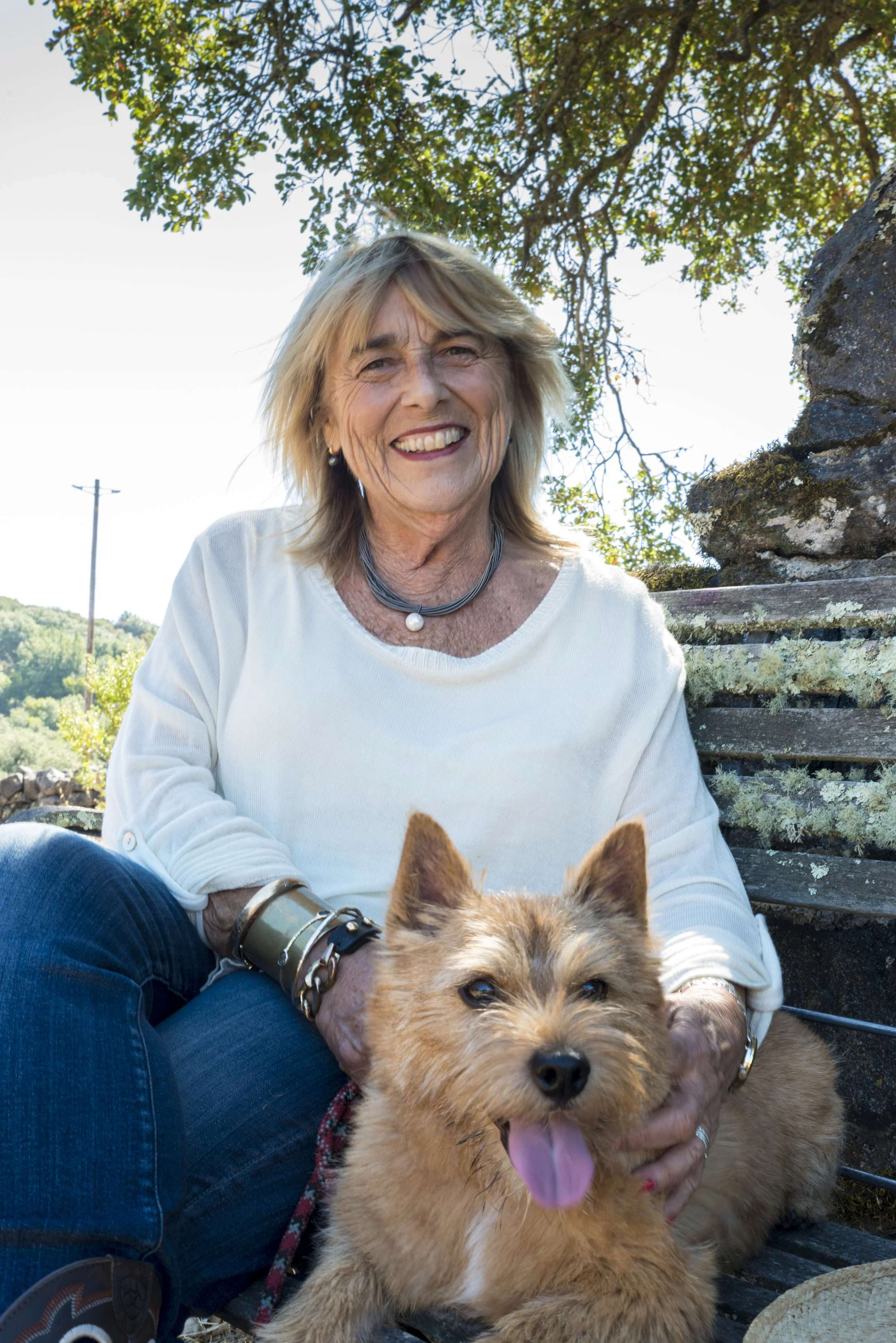 A smiling older woman with blonde hair sitting outdoors on a bench, holding a fluffy, tan-colored dog with its tongue out. The background shows trees, a clear sky, and a power line.
