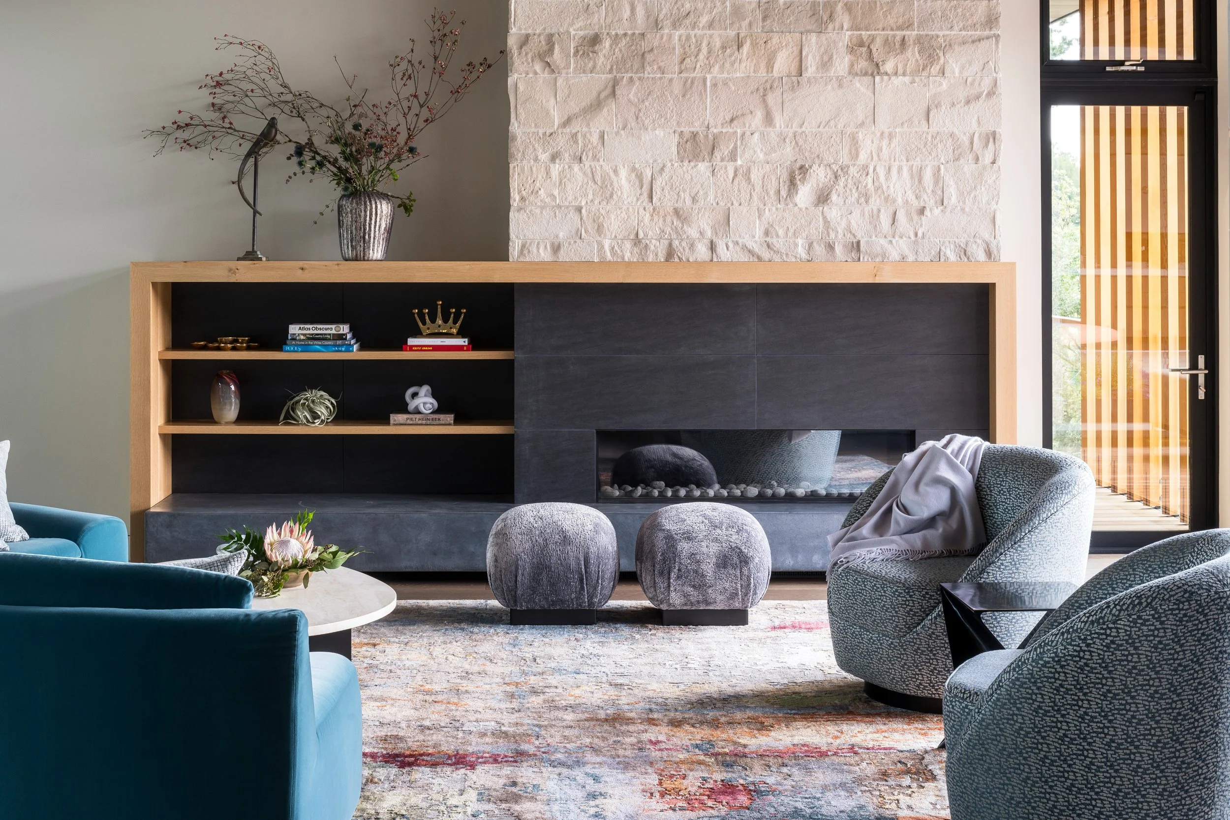 Modern living room with a black fireplace, wooden shelf with books and decorative items, large window with vertical blinds, and a variety of upholstered chairs and a round coffee table with flowers.