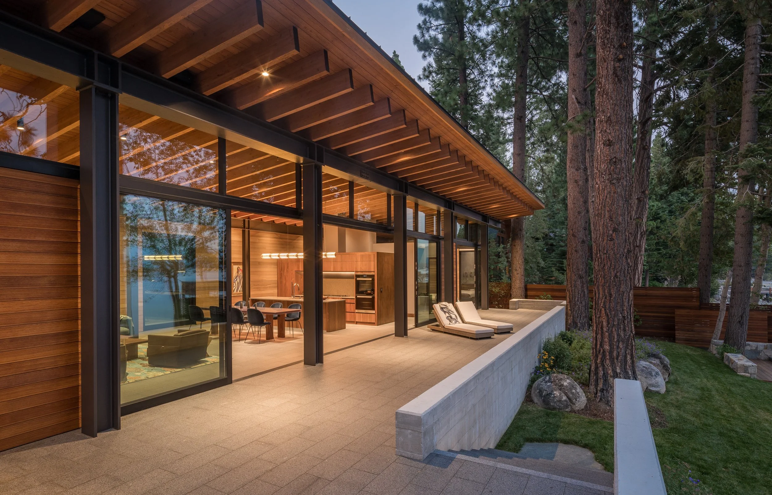 Modern house with large glass windows and a wooden roof, located in a wooded area with tall trees. The outdoor patio features lounge chairs and a dining area, illuminated by warm lighting at dusk.